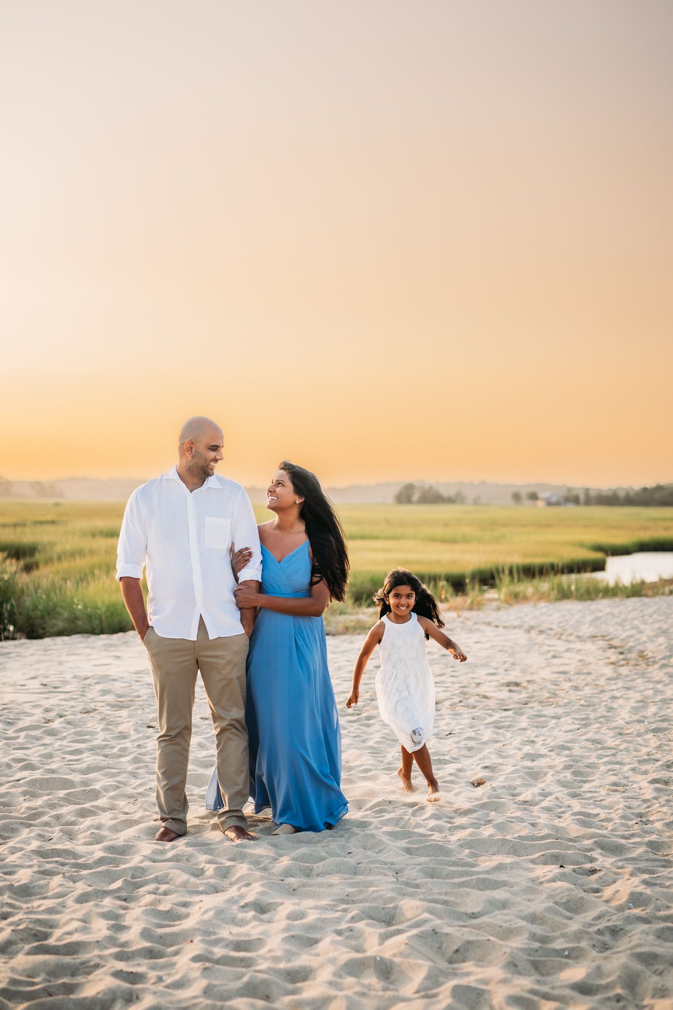 Family of three enjoying a sunset beach walk, mother in blue dress, father in white shirt, young daughter in white dress.