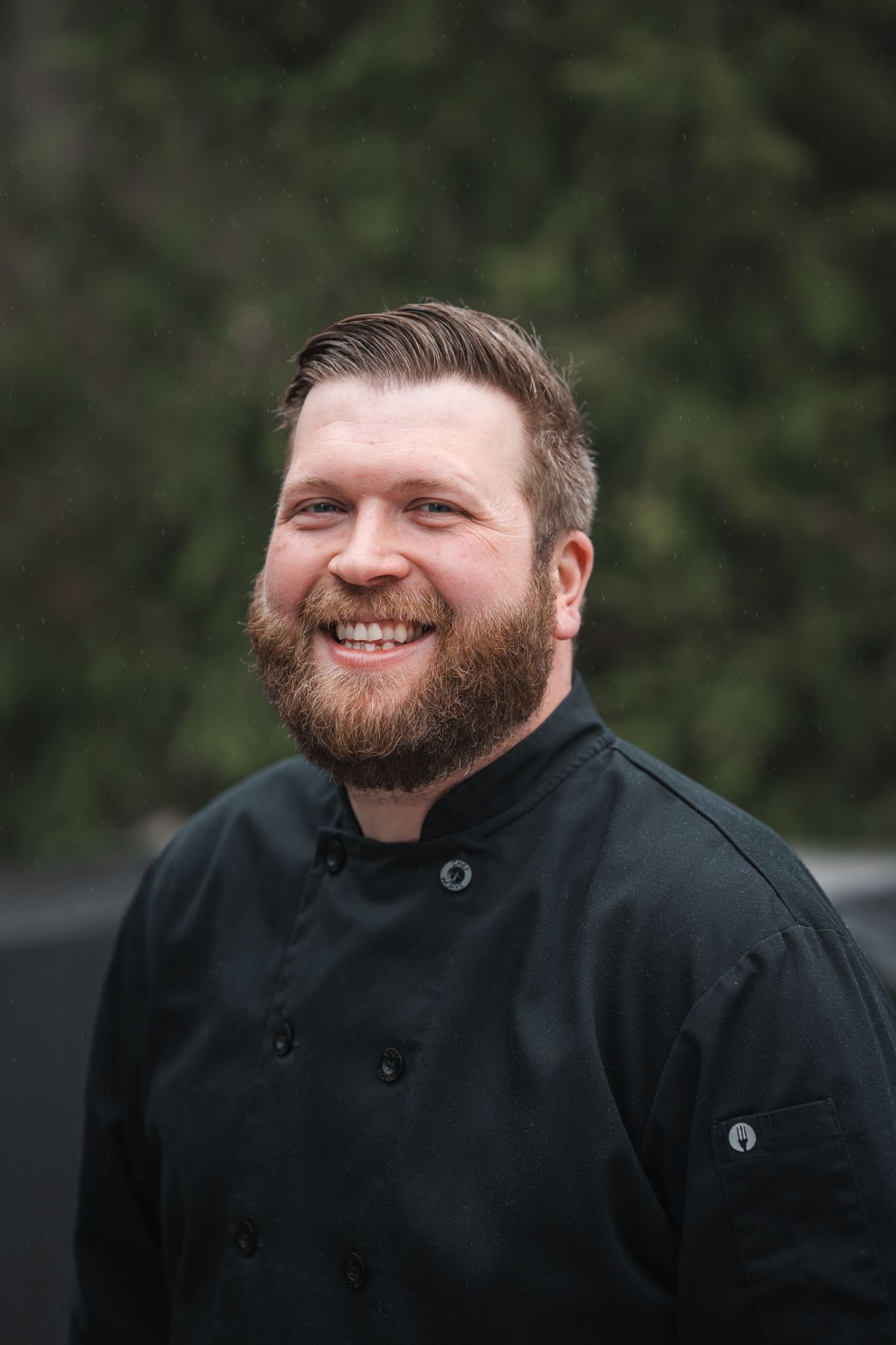 Smiling male chef with beard wearing black chef coat outdoors with green trees in background.