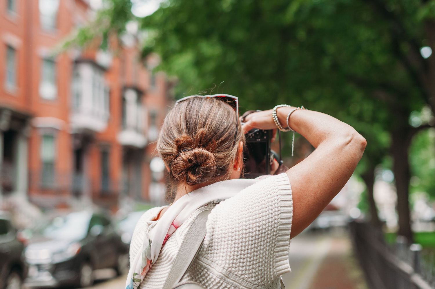 A person wearing a cream sweater stands with their back turned on a tree-lined residential street.