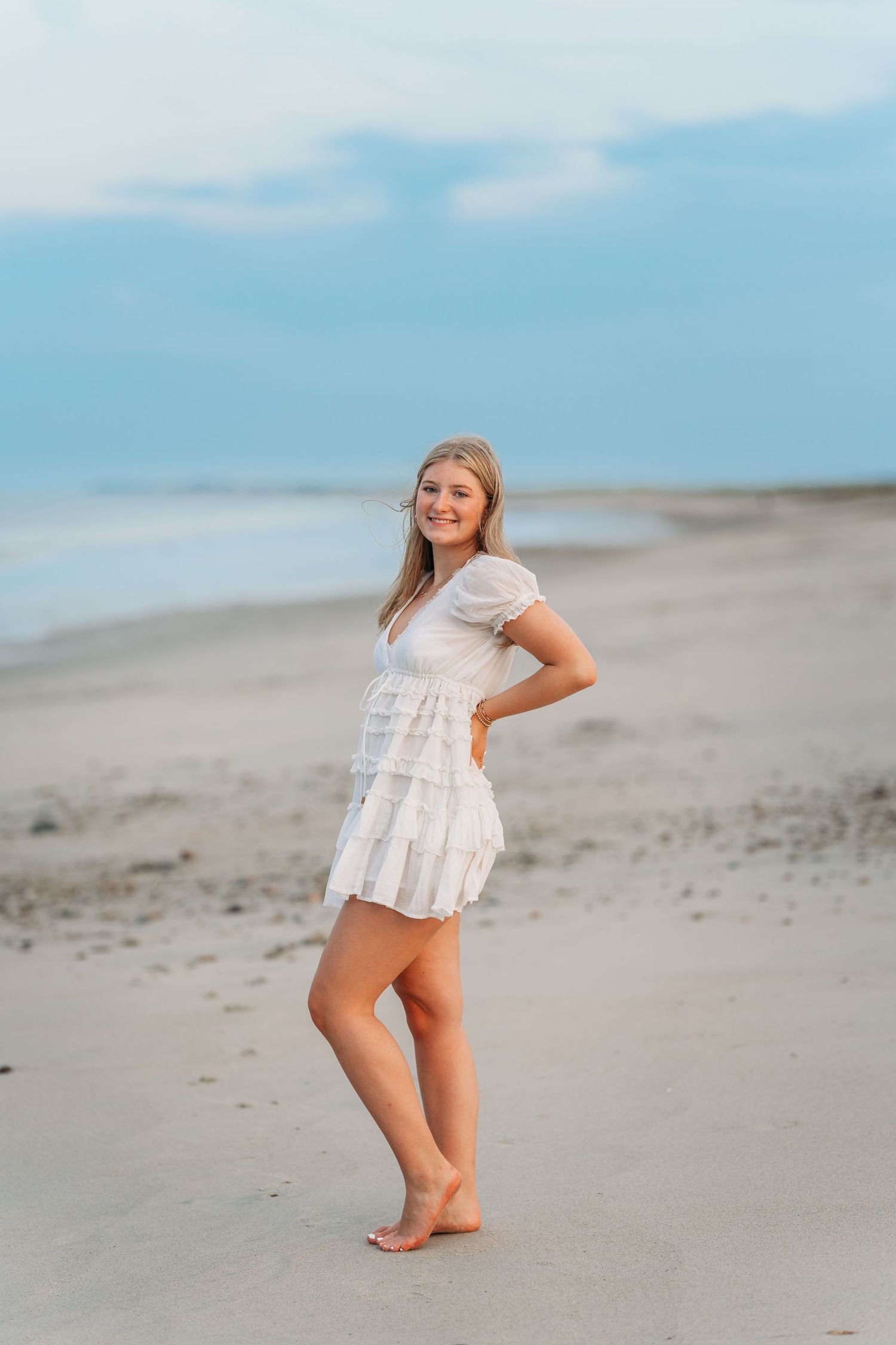 Young woman in white ruffled mini dress posing barefoot on sandy beach with ocean and cloudy blue sky.
