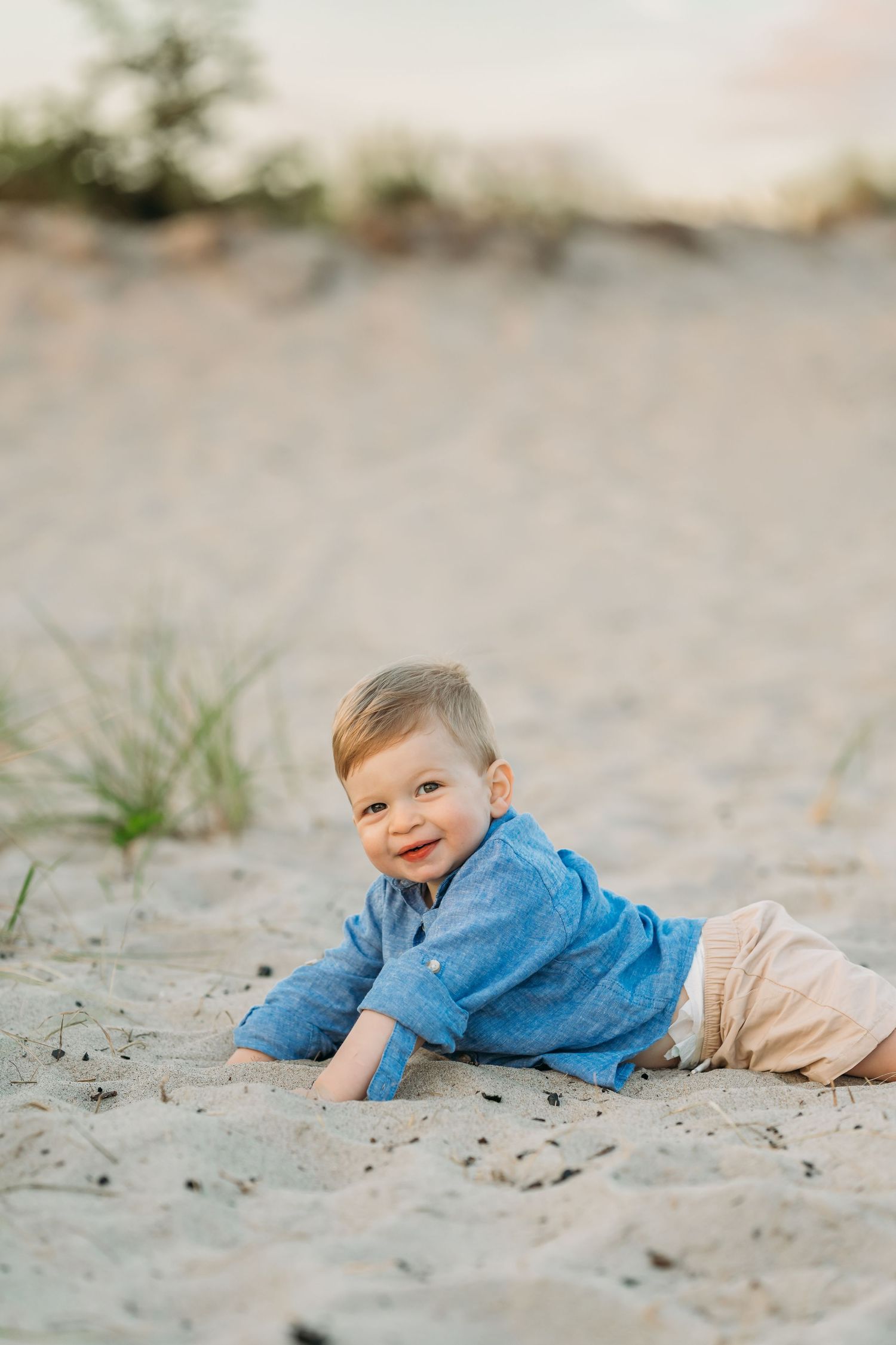 A small child wearing a blue shirt and beige pants crawls on the sand during a beach photo session.