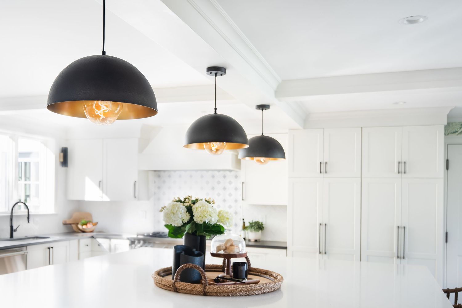 Modern white kitchen with black dome pendant lights, marble countertops, and white cabinetry features a round wooden tray display.