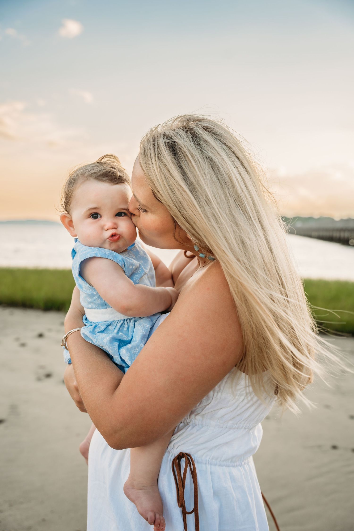 A tender moment captured between parent and child in white clothing against a serene beach backdrop at sunset.