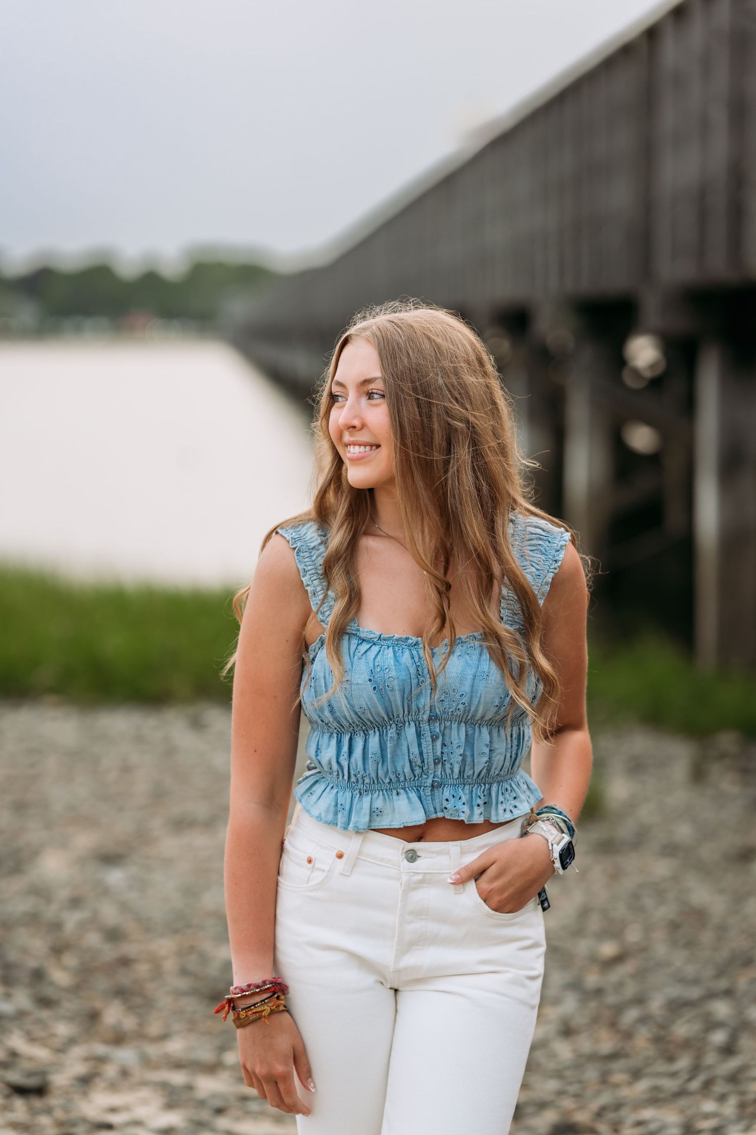 A person in a ruffled blue denim top and white jeans poses outdoors on a rocky beach.