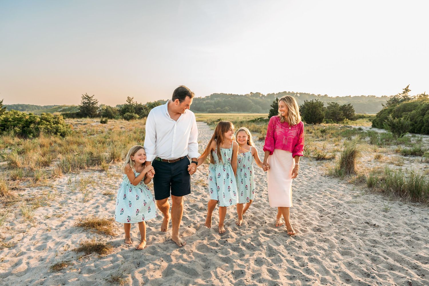 A family walks together along a sandy beach path during golden hour with coastal vegetation in the background.