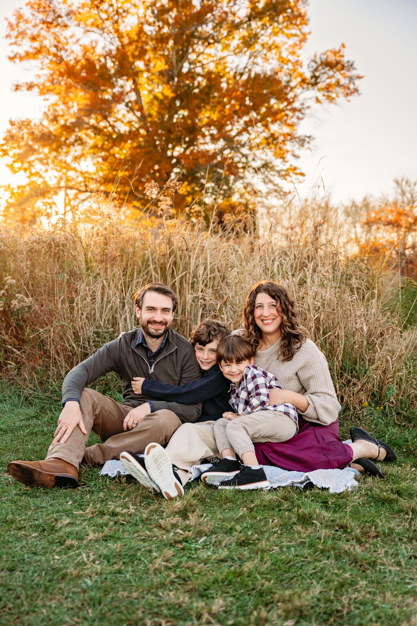 Family of four sitting on a blanket in a field during golden hour fall sunset with colorful autumn tree backdrop.