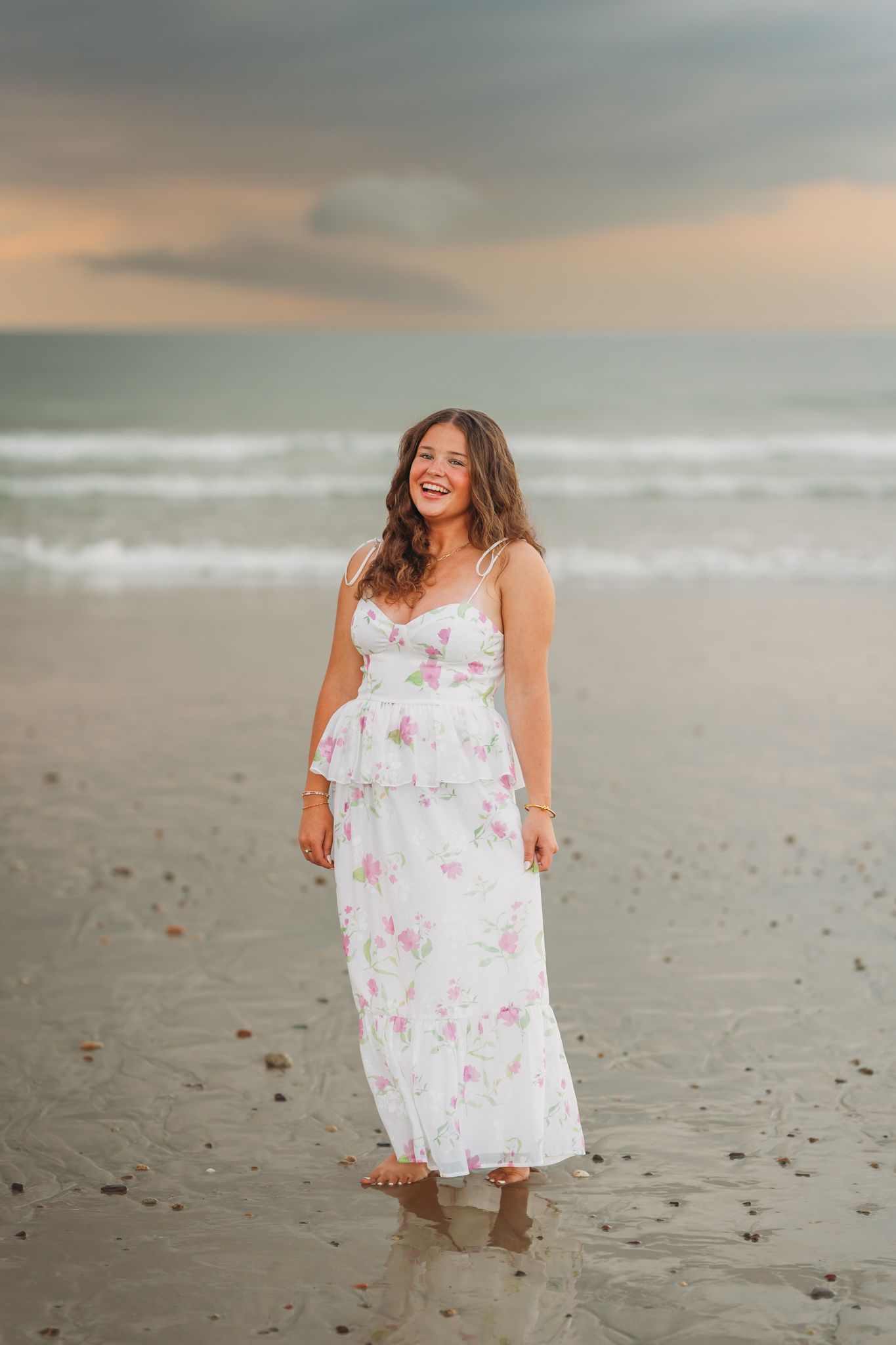 A smiling young woman in a white floral maxi dress stands barefoot on a sandy beach at sunset.
