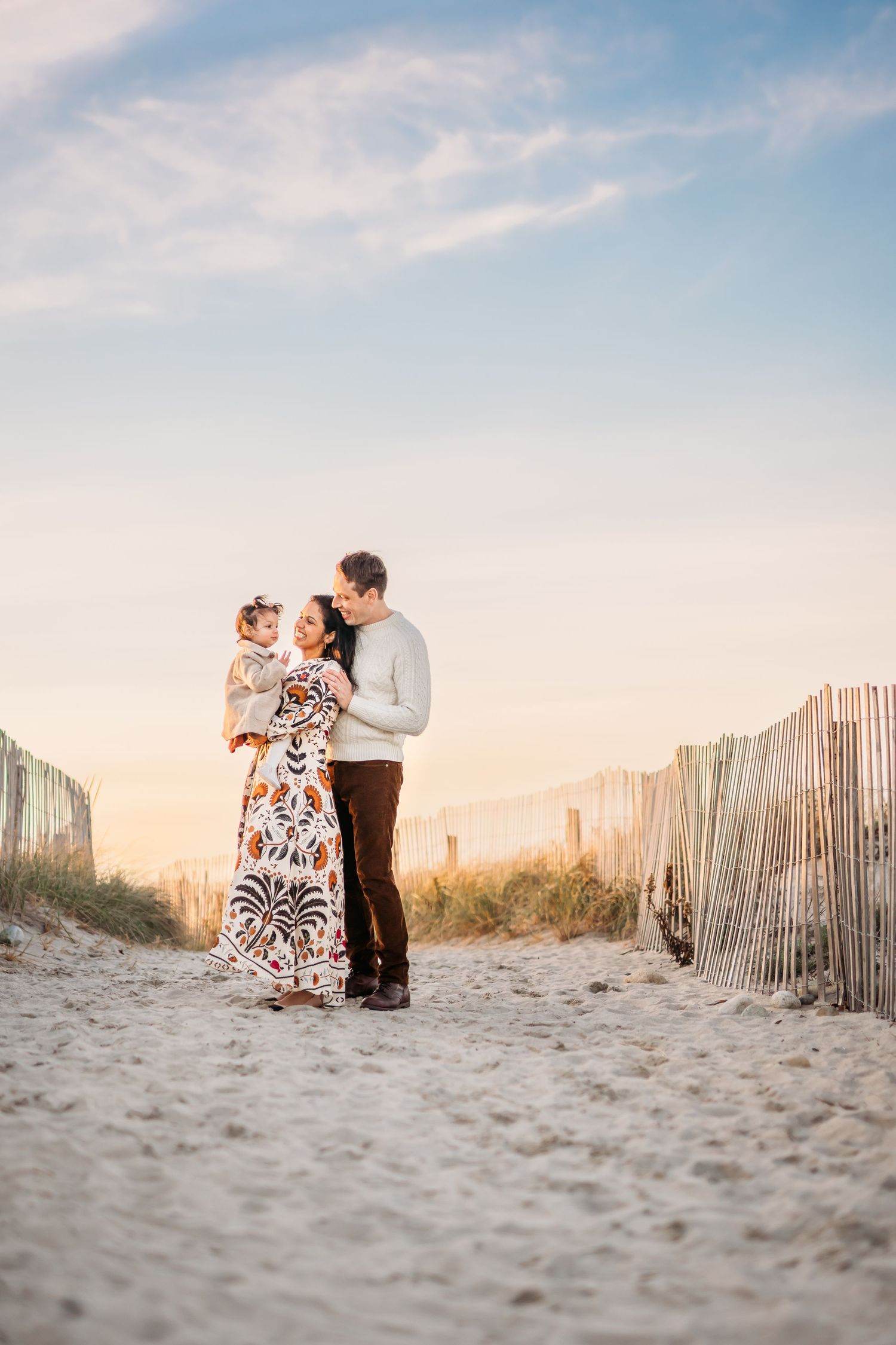 Beautiful family moment captured on sandy beach path at sunset with wooden fence and warm sunlit sky in background.