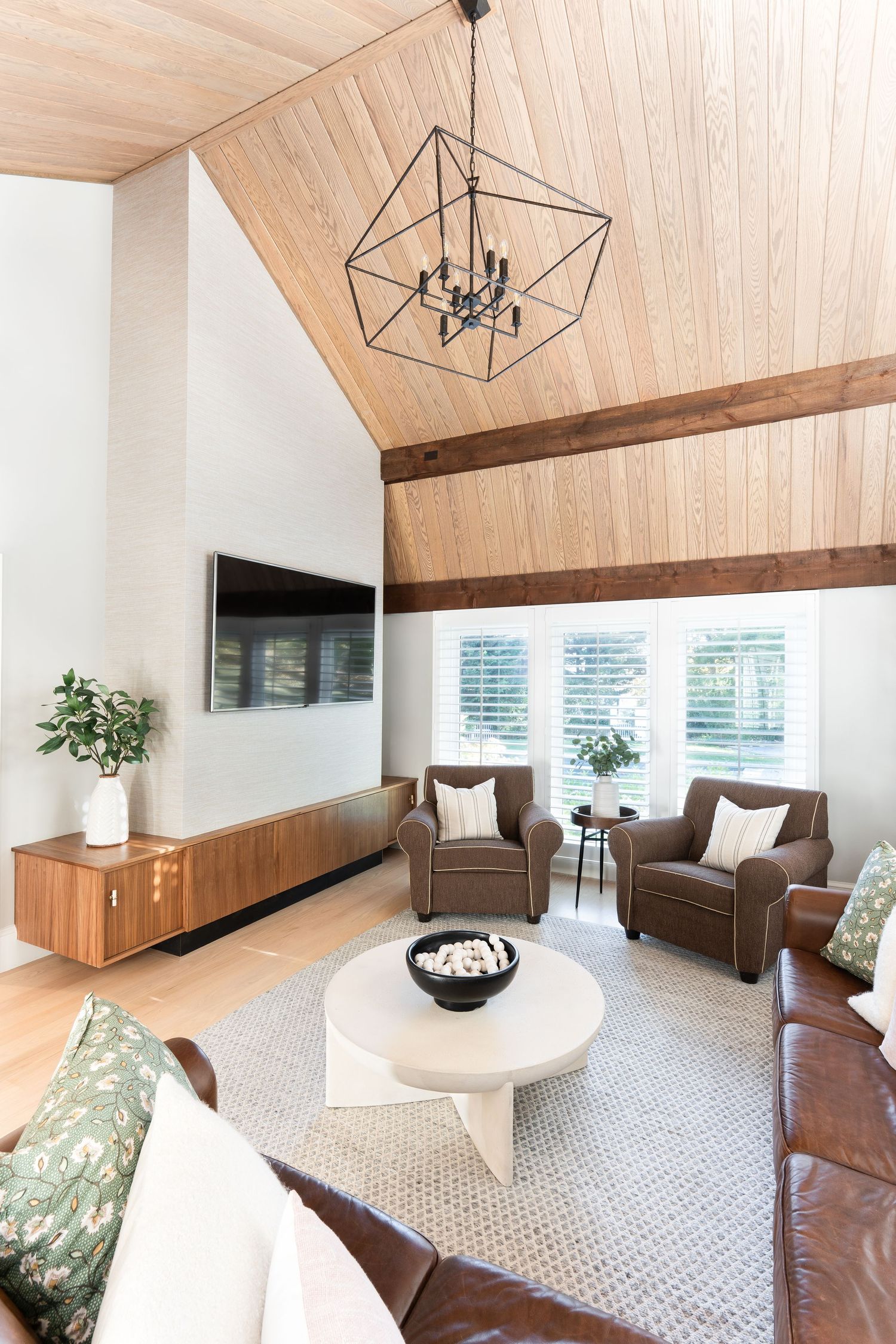 Modern living room with vaulted wood ceiling, geometric chandelier, brown leather chairs and white curved sofa on patterned carpet.
