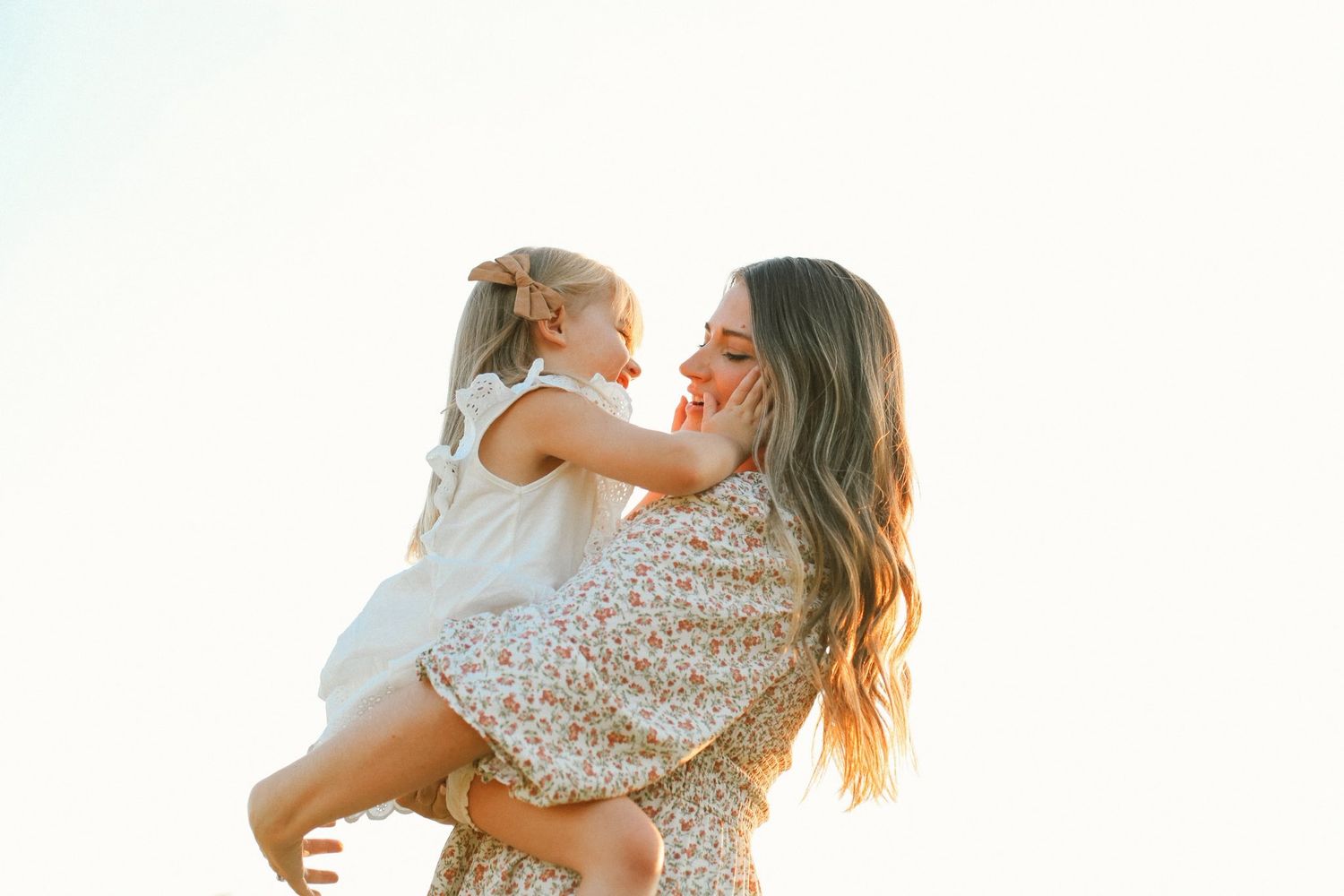 A parent and child share a joyful moment together in golden sunlight while wearing floral dresses.