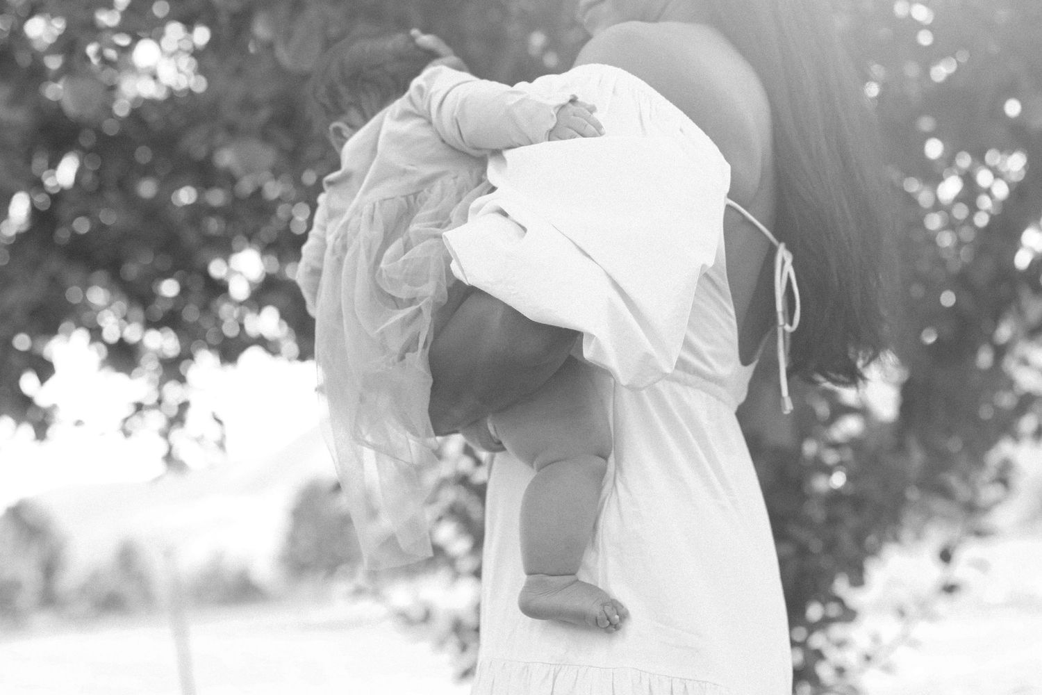 Black and white image of someone holding a child up in the sunlight against trees.