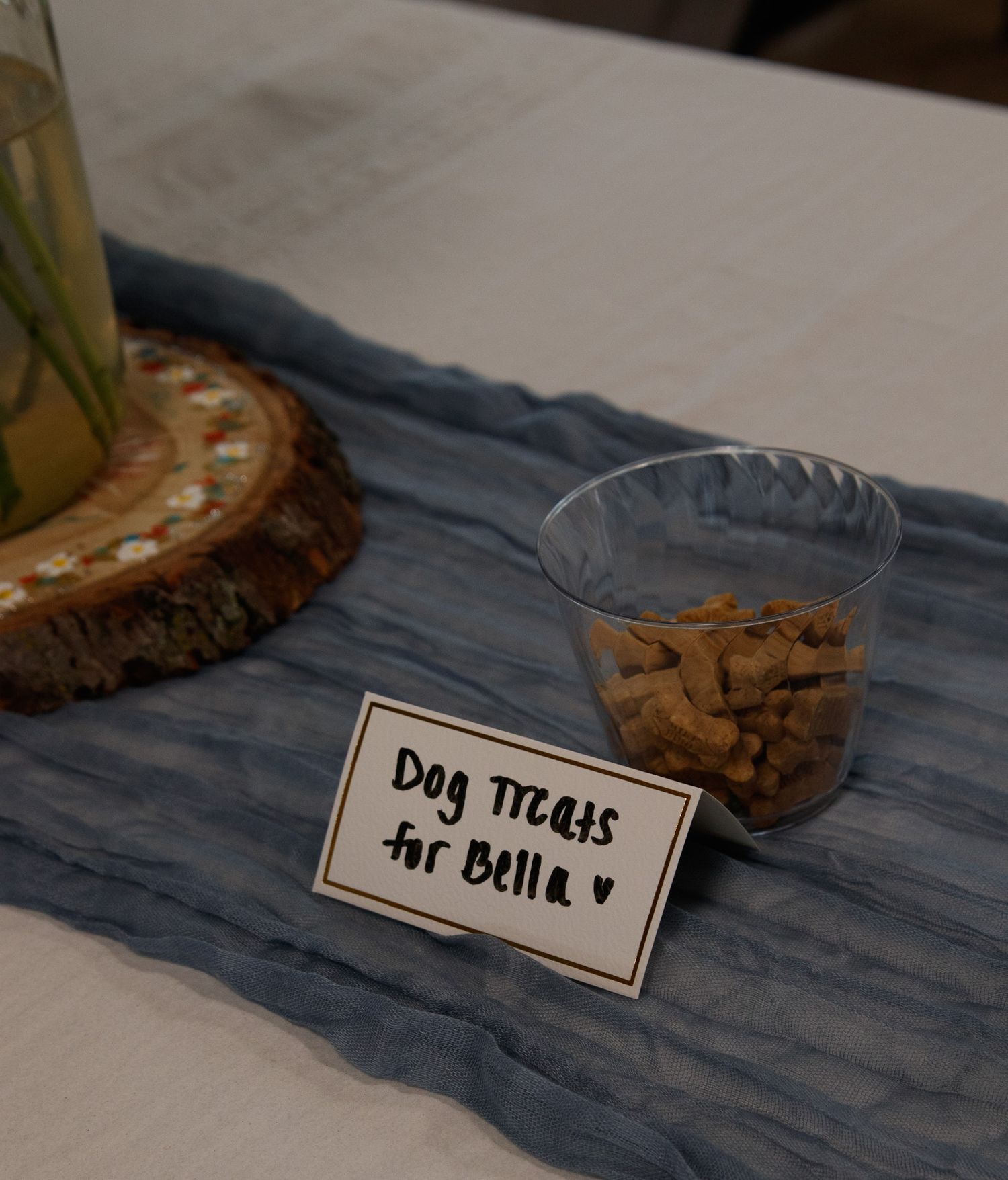 A glass ramekin of dog treats sitting next to a small hand-lettered sign that says 'Dog treats for Bella' on a blue napkin.