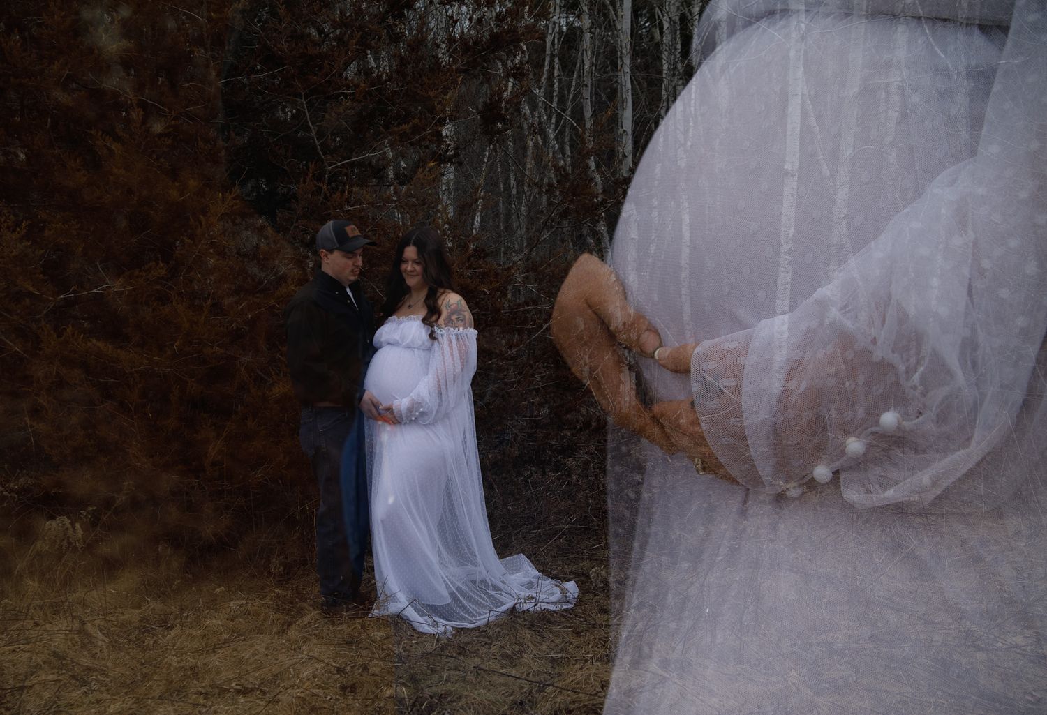Couple embracing outdoors during maternity photoshoot, woman in flowing white gown with pregnant belly.