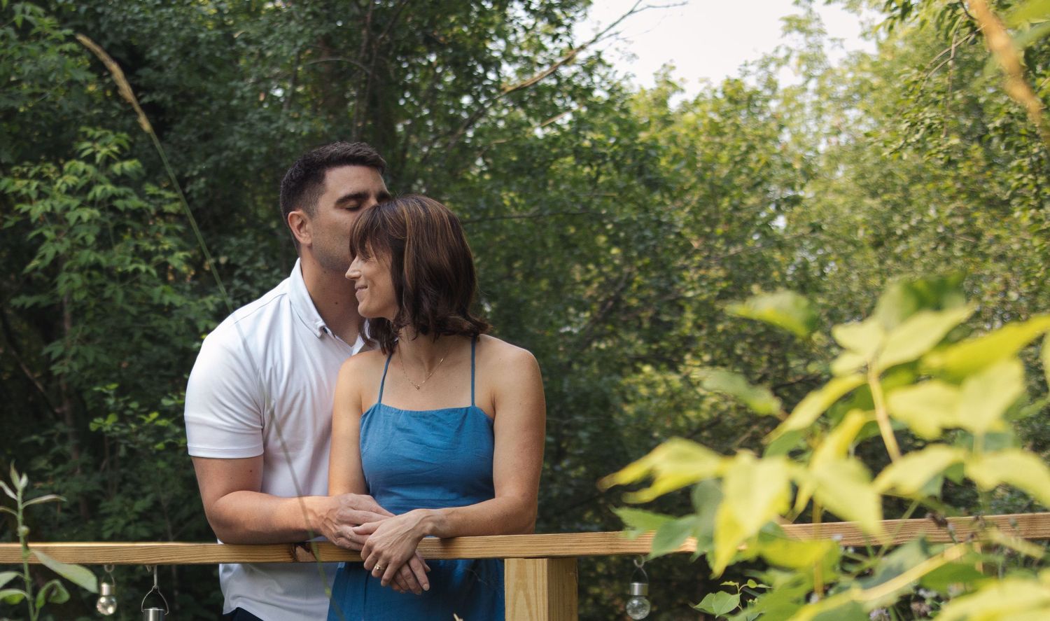 Romantic couple embraces in a lush green garden setting with sunlight filtering through the trees.