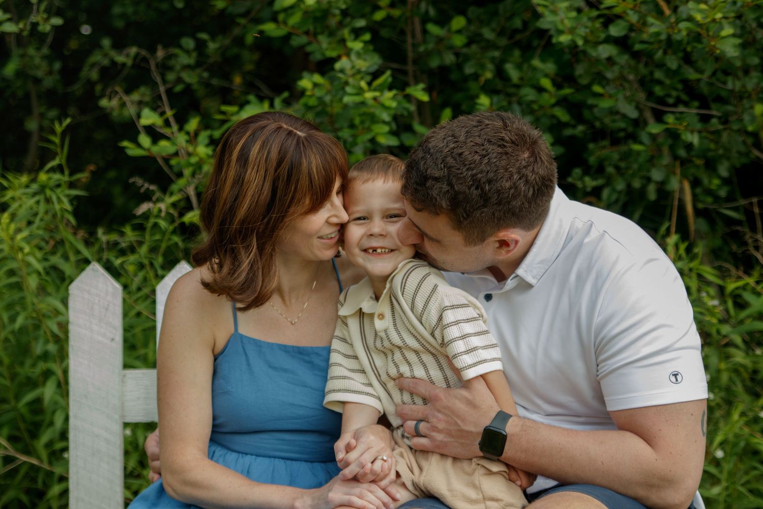 Happy family embraces while sitting together on white fence during sunset photo shoot.