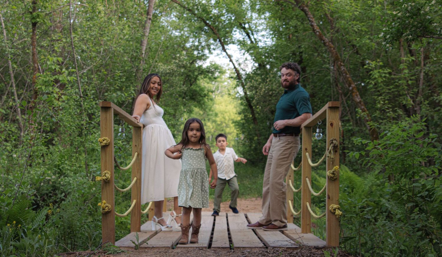 A family stands together smiling on a wooden bridge surrounded by lush green trees and foliage on a sunny day.