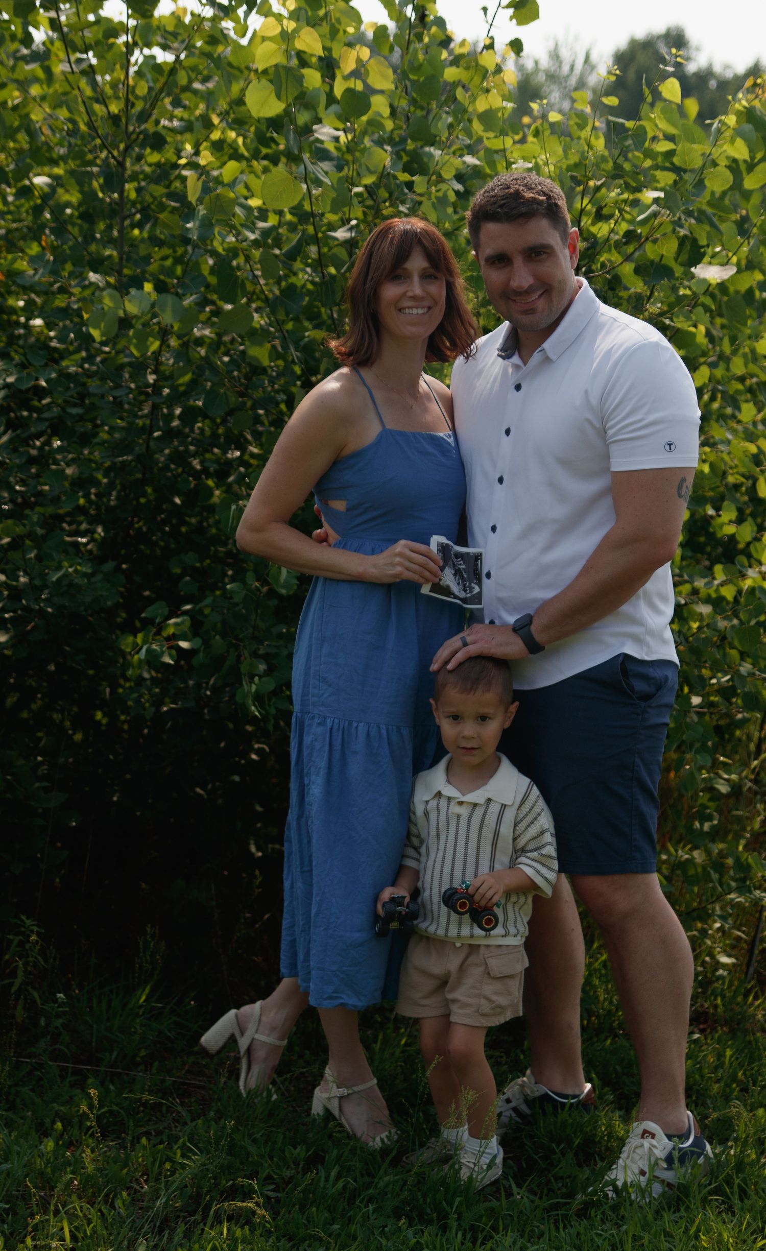 A family poses together outdoors among lush green foliage on a sunny summer day.