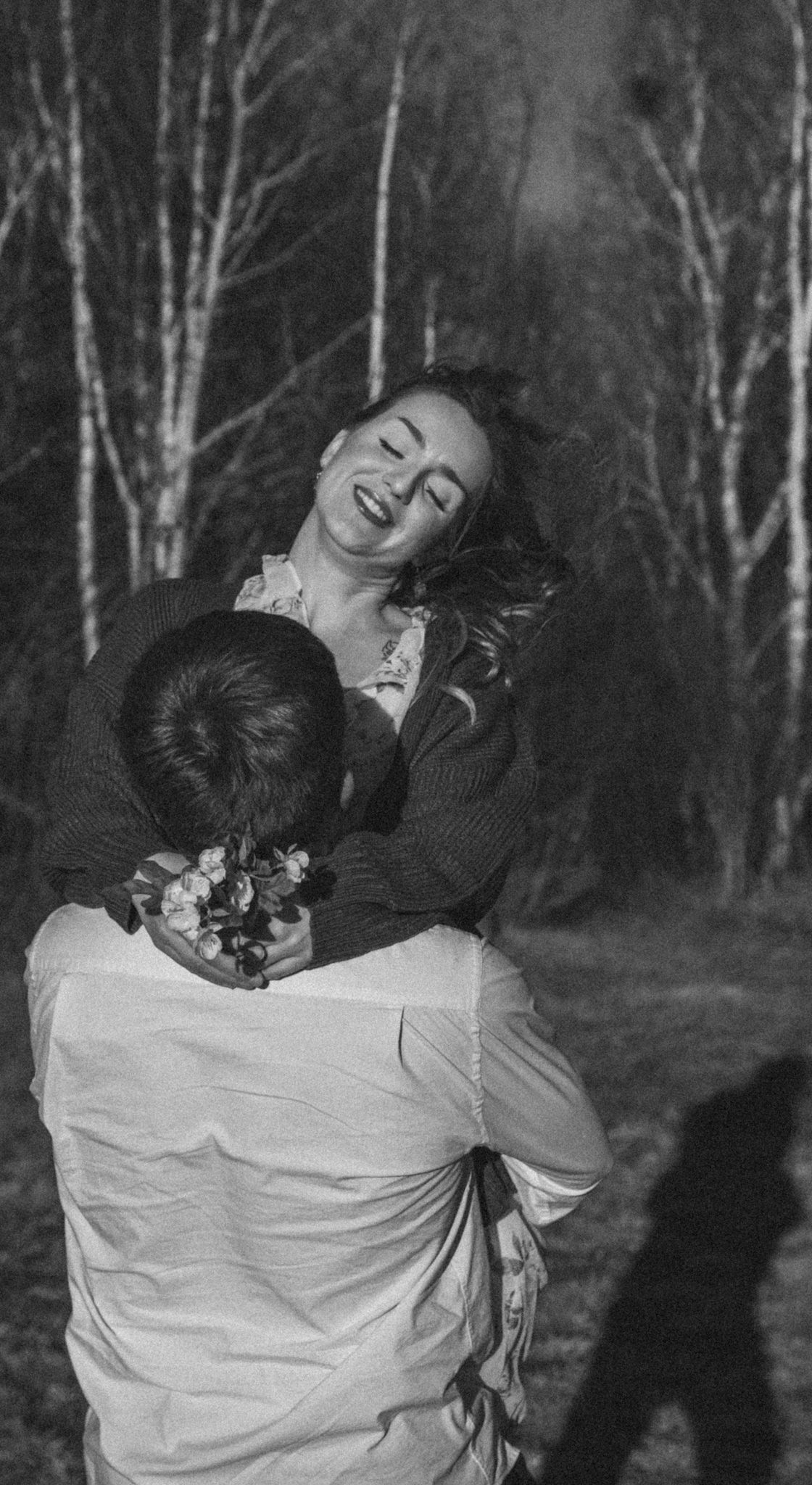 Black and white photo of a joyful woman being lifted by a man holding flowers in a birch forest at night.
