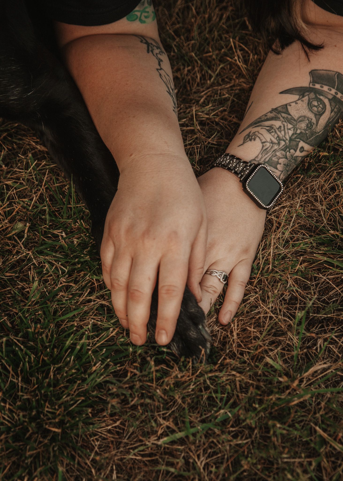 Tattooed woman wearing an Apple Watch and silver ring gently holds a black dog's paw on green grass.