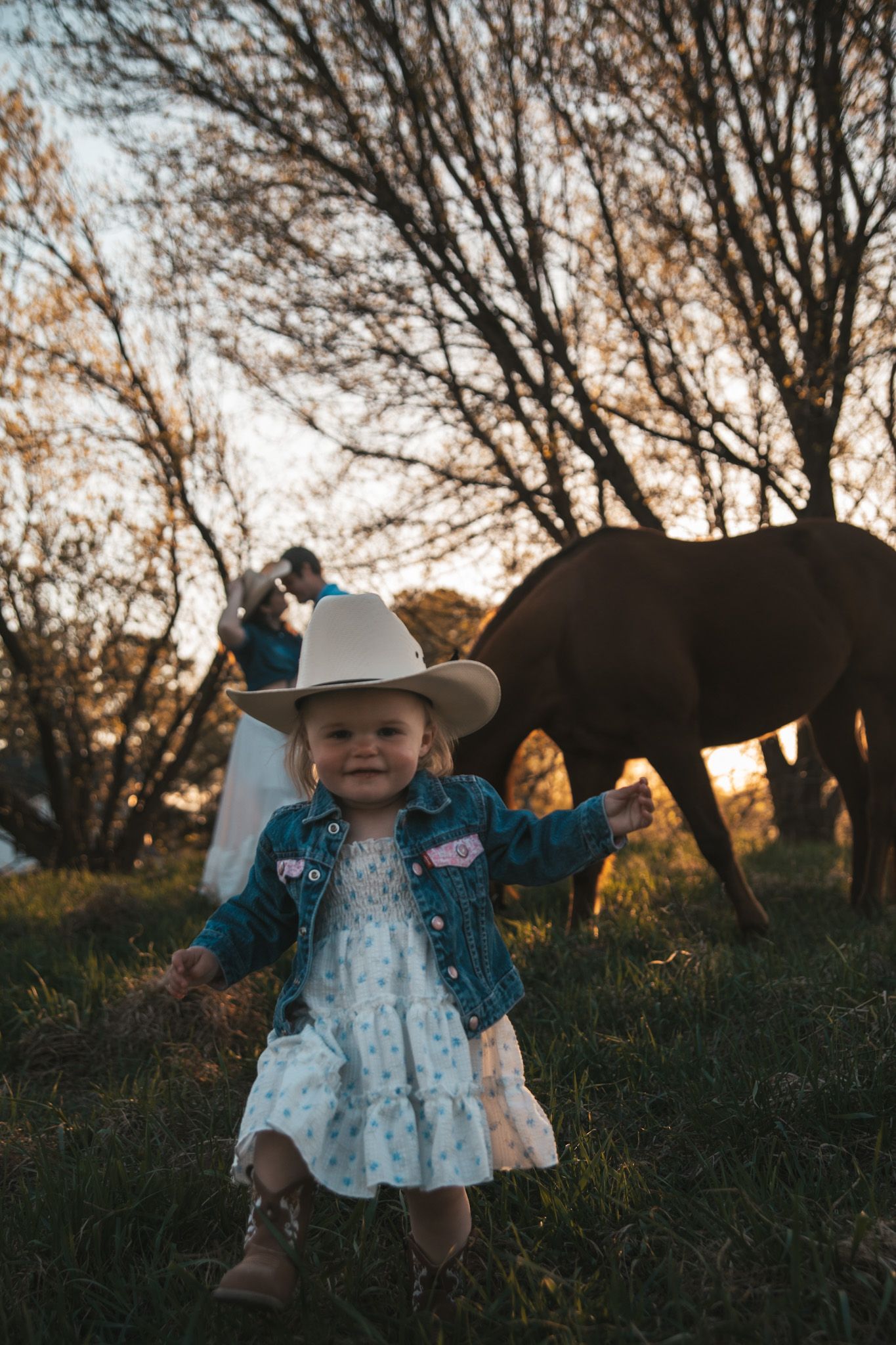 Smiling toddler in oversized cowboy hat and denim jacket poses with horse and parents at golden hour.