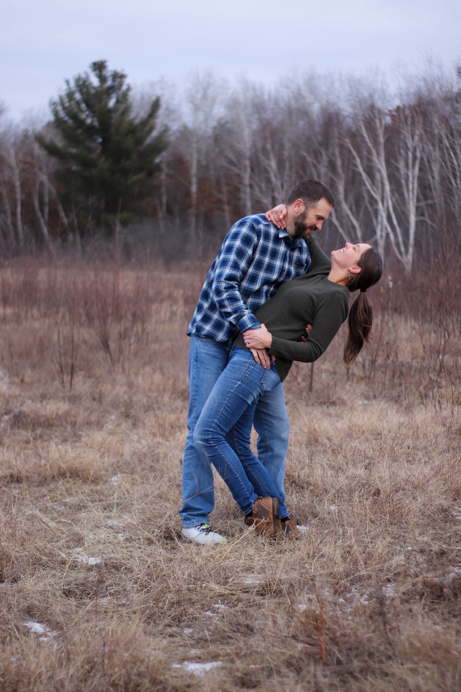 A romantic couple embraces in a winter field with bare trees in the background.
