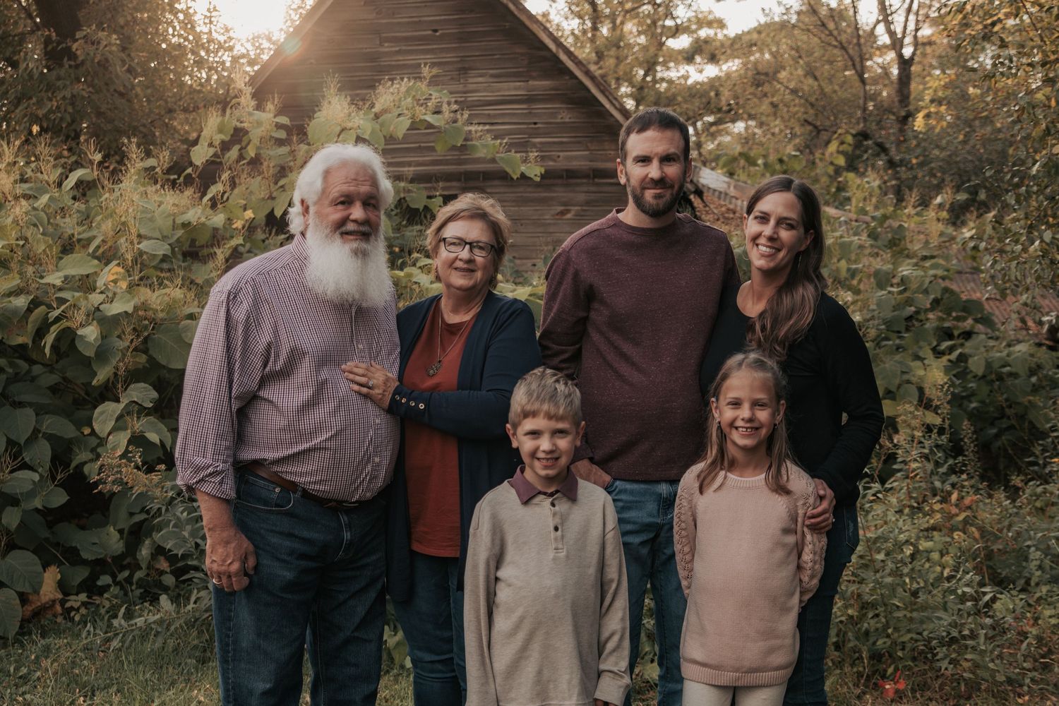 A group of people pose together outdoors during a fall photo session with trees and natural lighting in the background.
