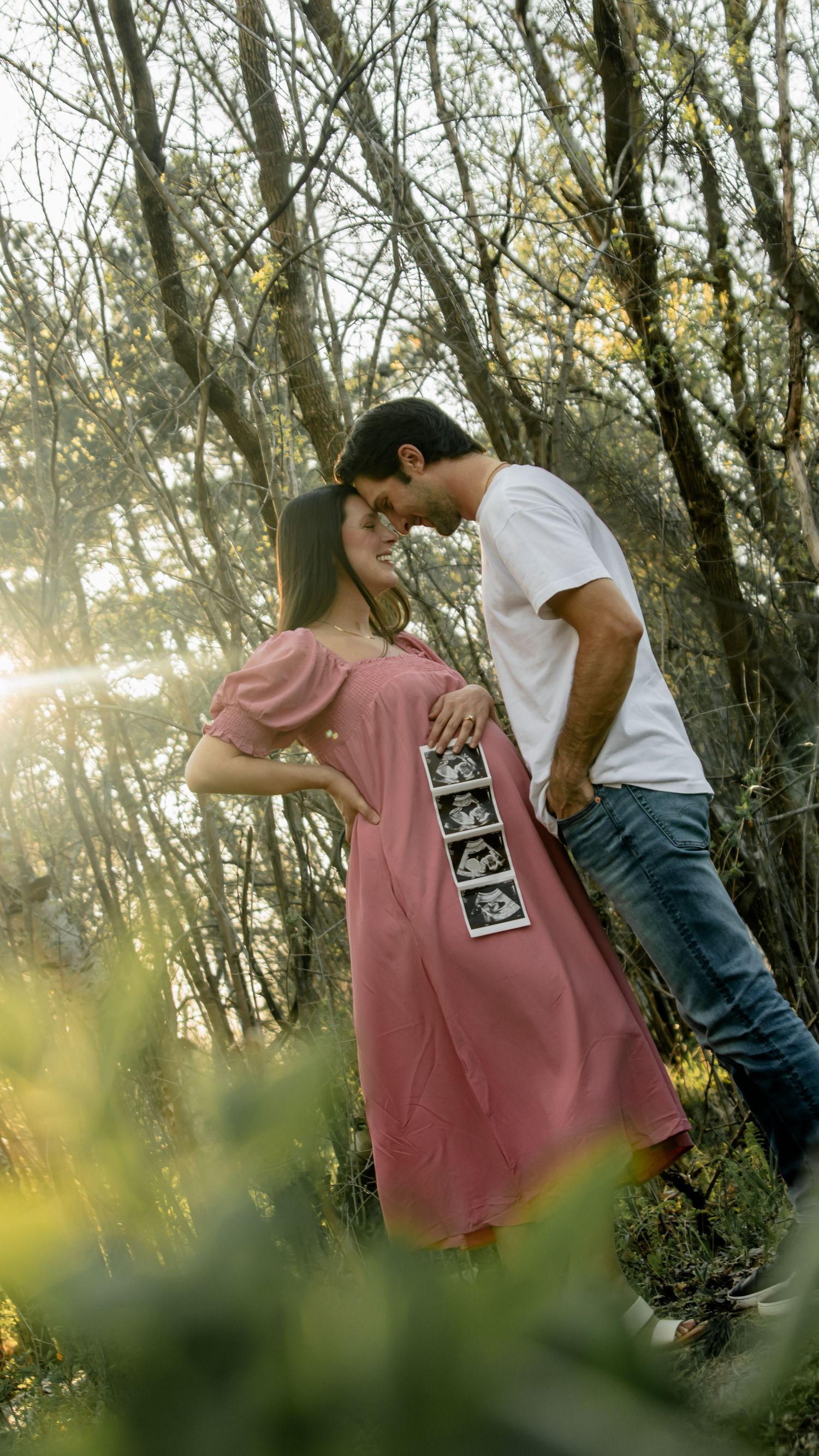 A couple shares an intimate sunset moment in a forest while holding ultrasound photos showing their pregnancy announcement.