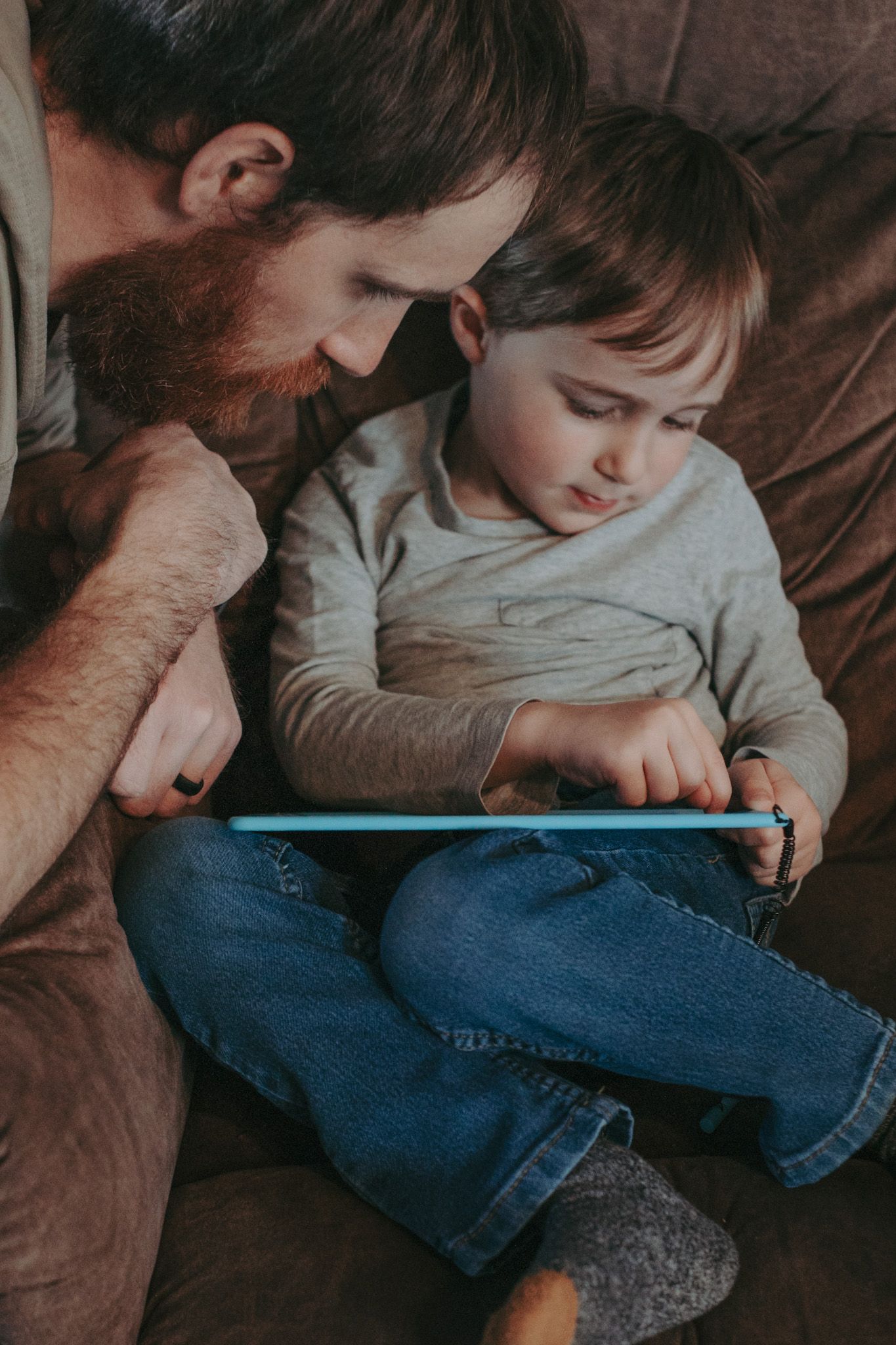 Bearded father and young toddler son sitting on couch together using a blue tablet device.