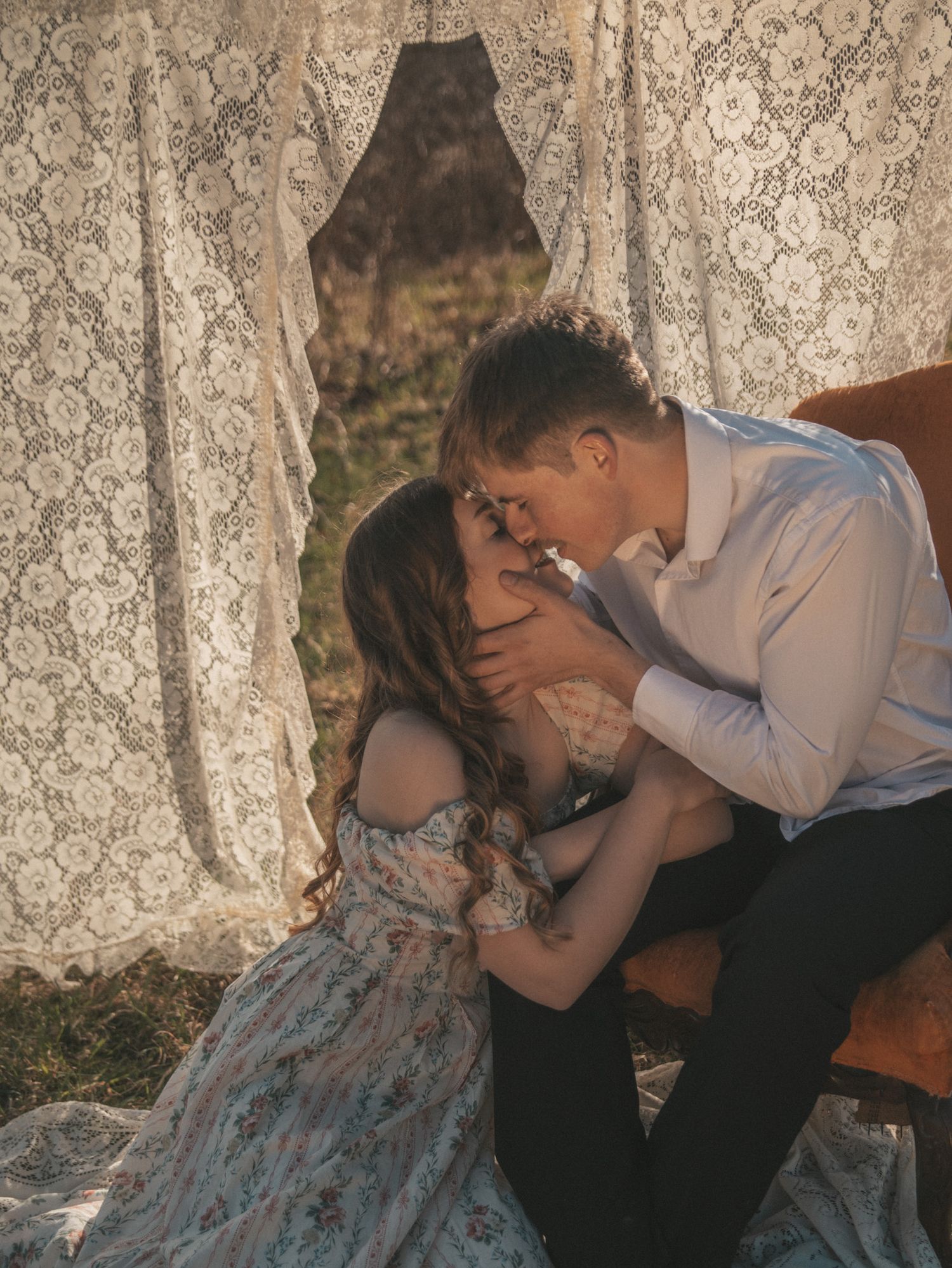 Romantic couple sharing an intimate moment under lace curtains outdoors in warm golden light.