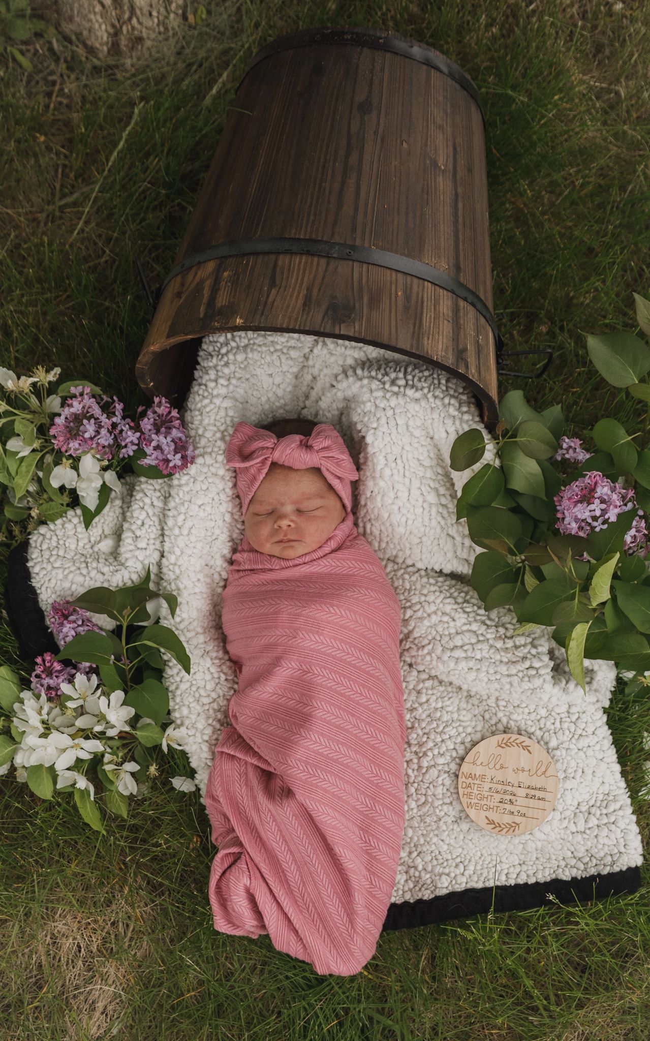 Newborn baby swaddled in pink wrap with bow headband, lying on fluffy white blanket beside wooden barrel and lilac flowers.