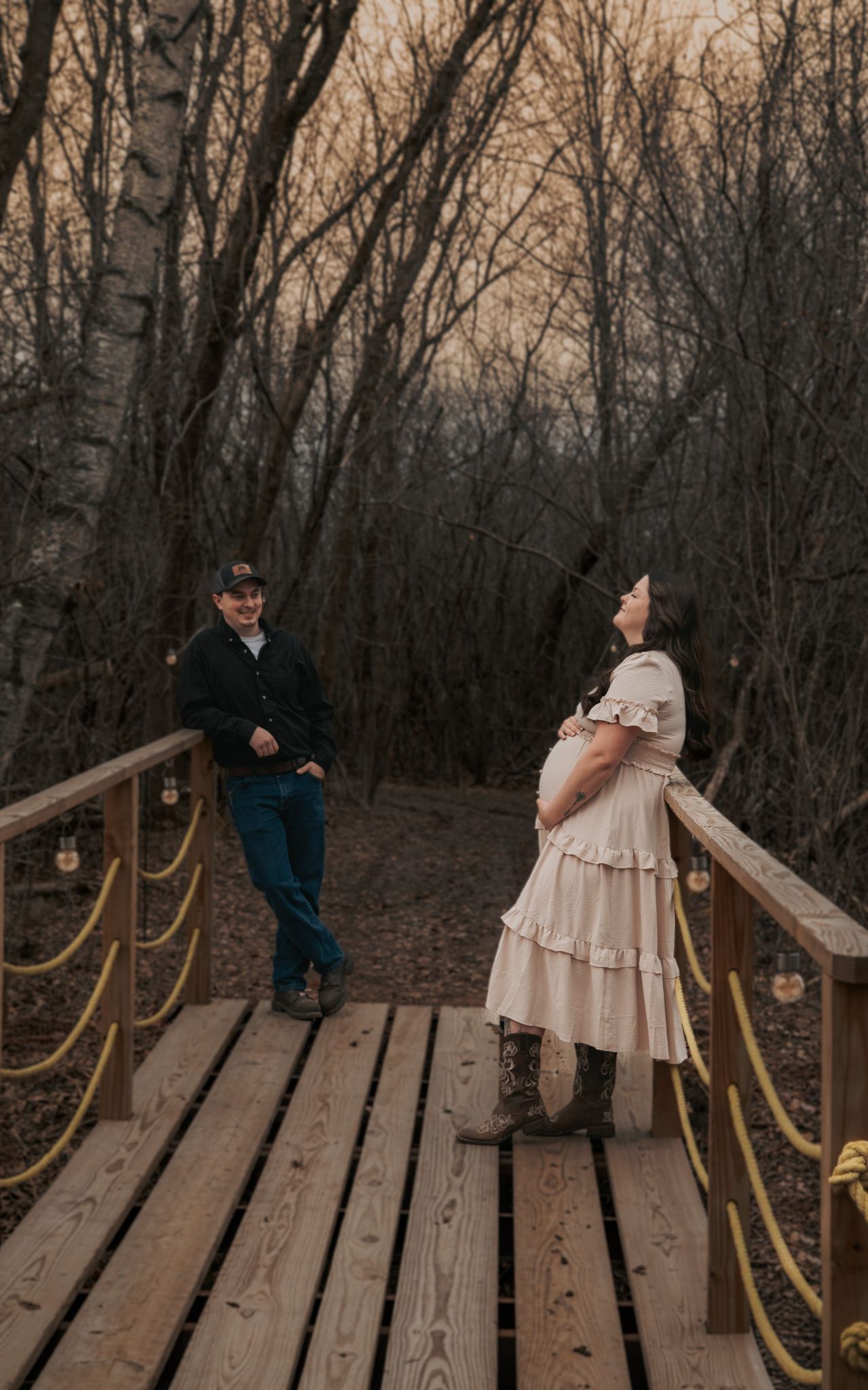 Pregnant woman in cream dress and cowboy boots stands on wooden bridge with partner among bare winter trees at dusk.
