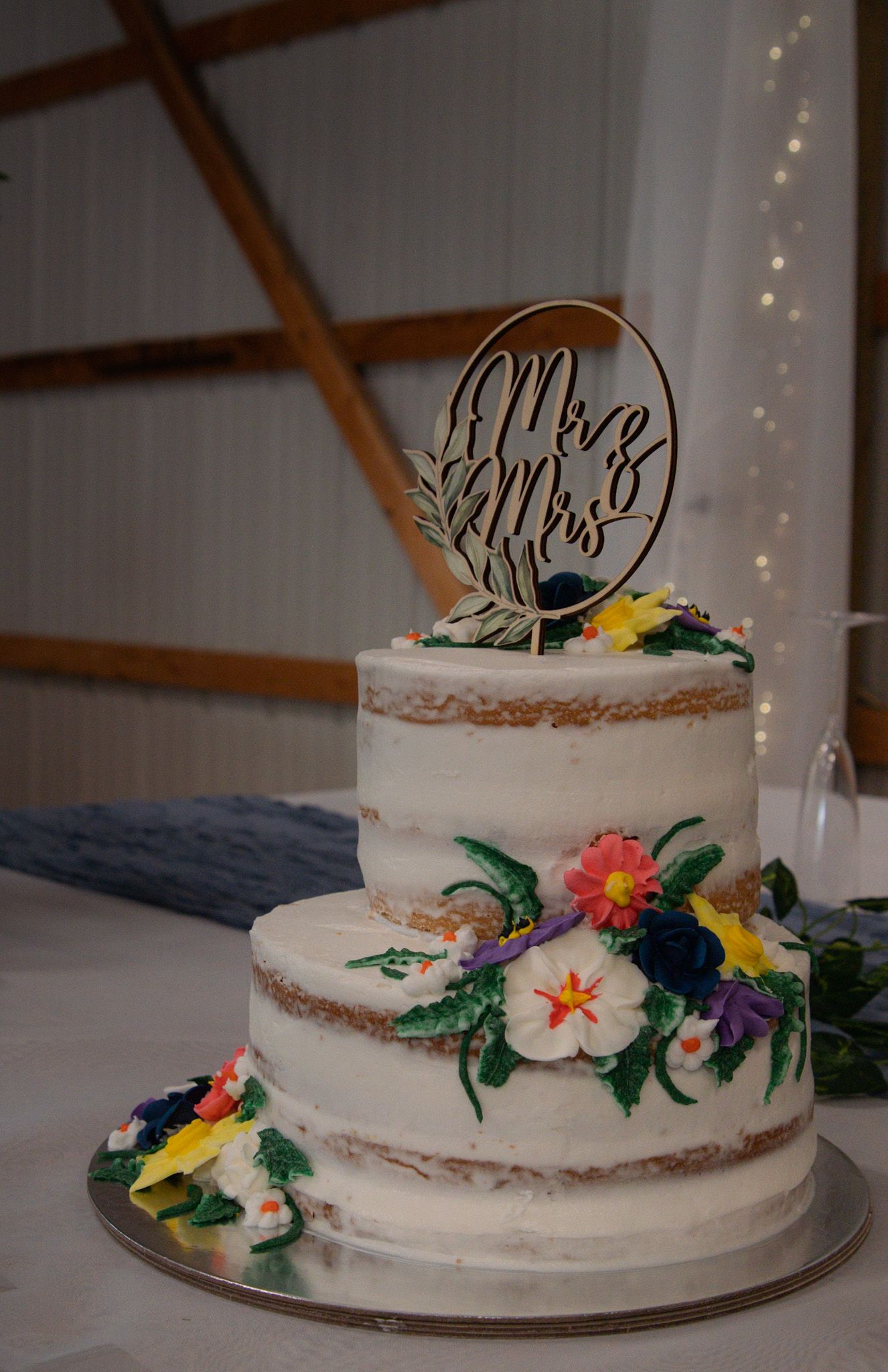 A white two-tiered wedding cake decorated with colorful tropical flowers and a decorative cake topper is displayed.