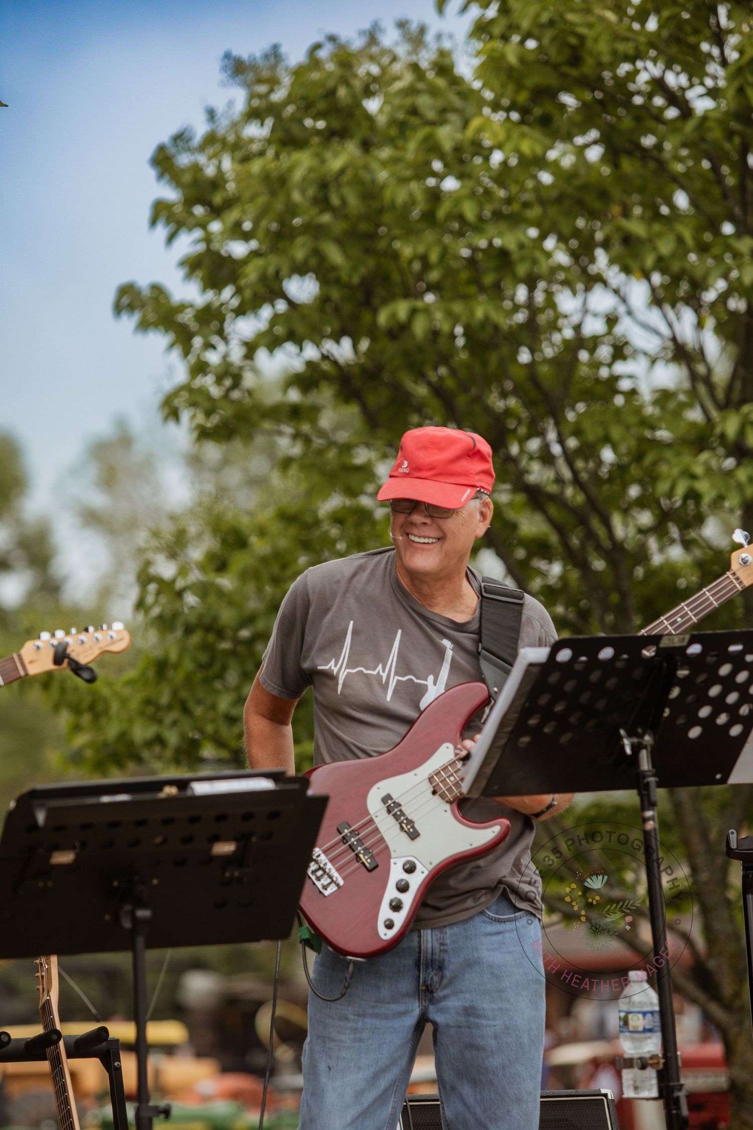 A guitarist in a red cap and gray shirt performs outdoors with a red bass guitar and music stand under green trees.
