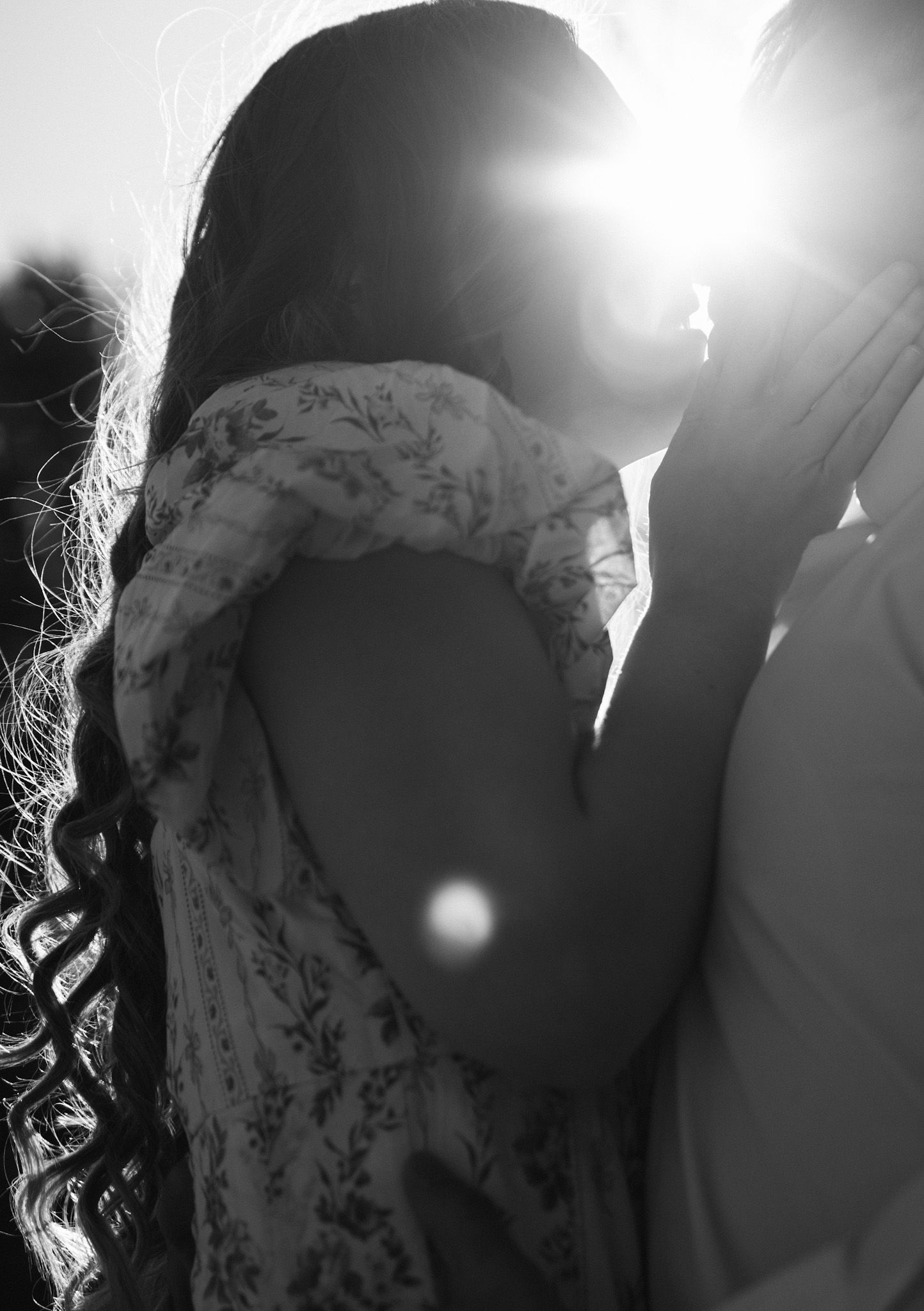 Black and white photo of a couple sharing a romantic kiss backlit by bright sunlight, creating a dreamy lens flare effect.