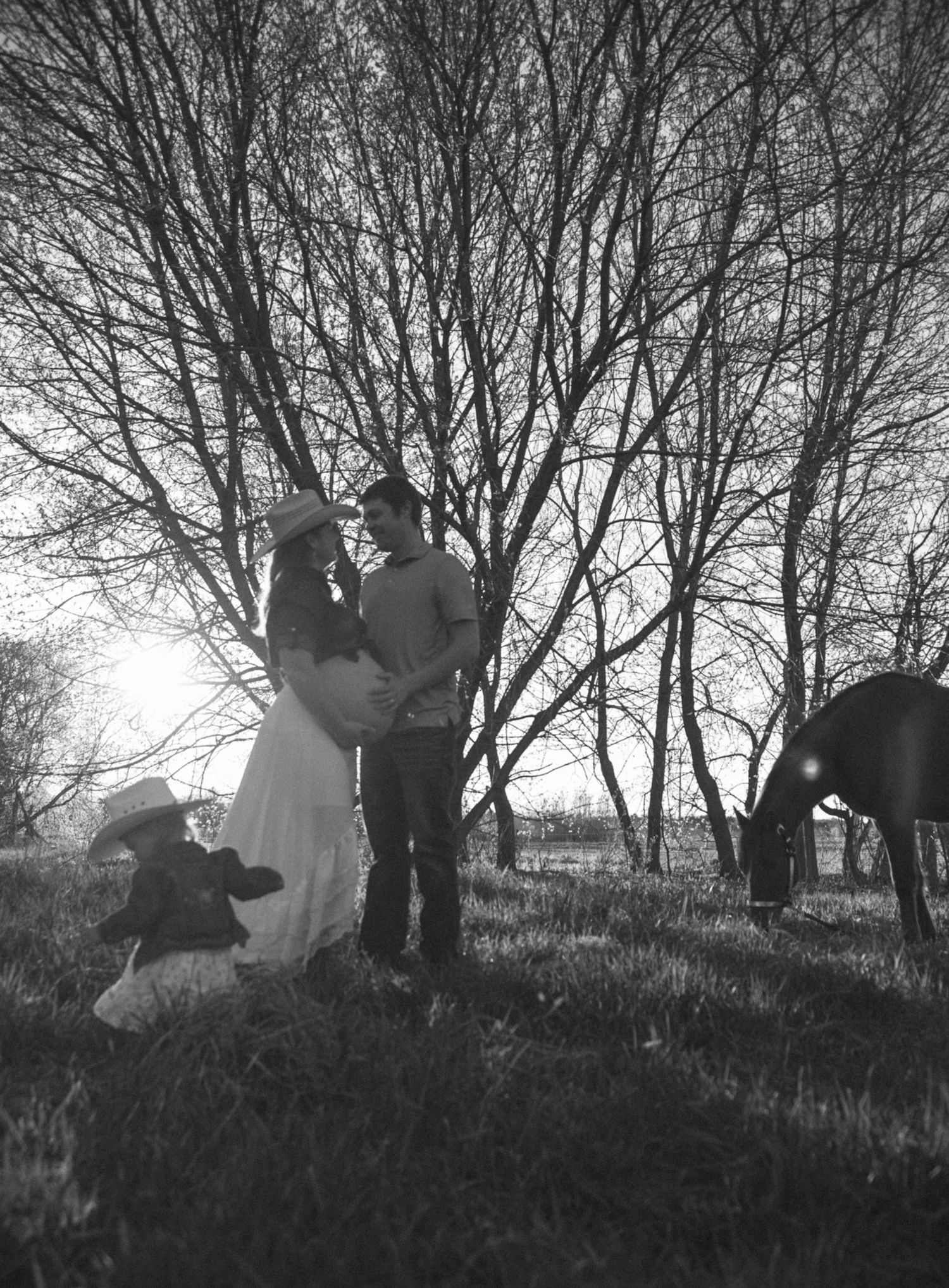 Black and white maternity photo of couple embracing outdoors with child and horse near bare trees.
