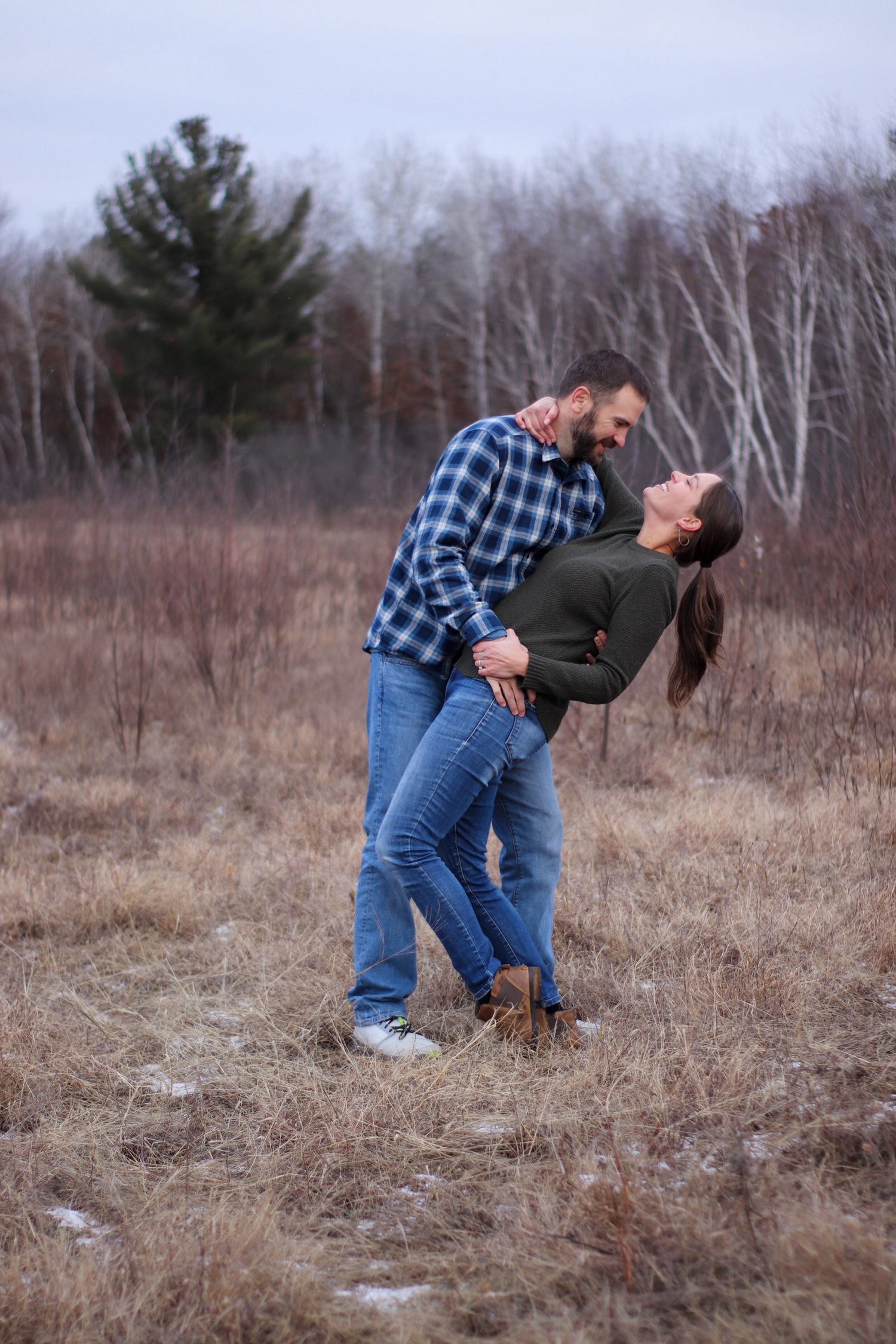 Romantic couple in casual attire sharing tender moment in rustic field with winter trees in background.