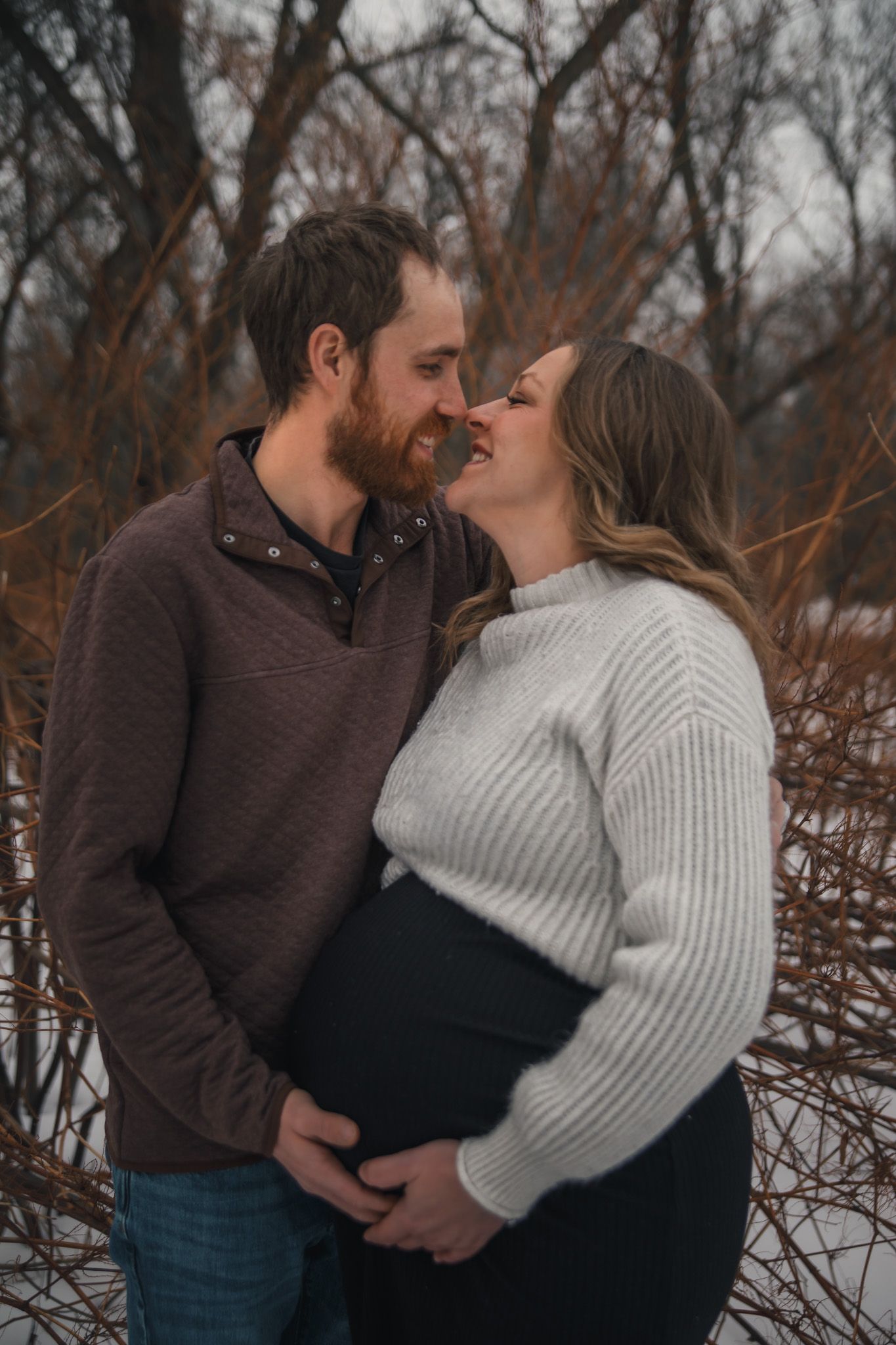 Romantic winter couple in brown and white sweaters embrace against bare trees in snowy landscape.
