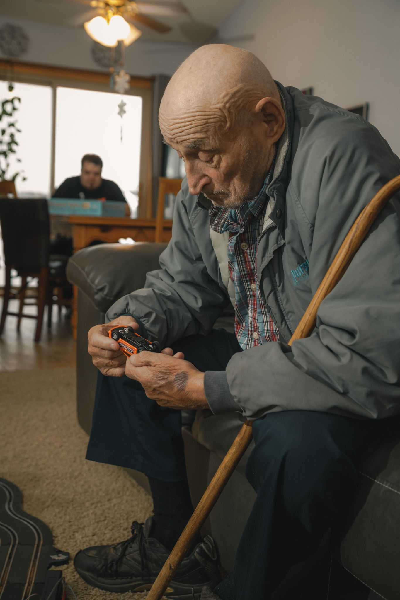Elderly bald man in gray jacket sits in chair holding a small device, leaning on wooden cane indoors.