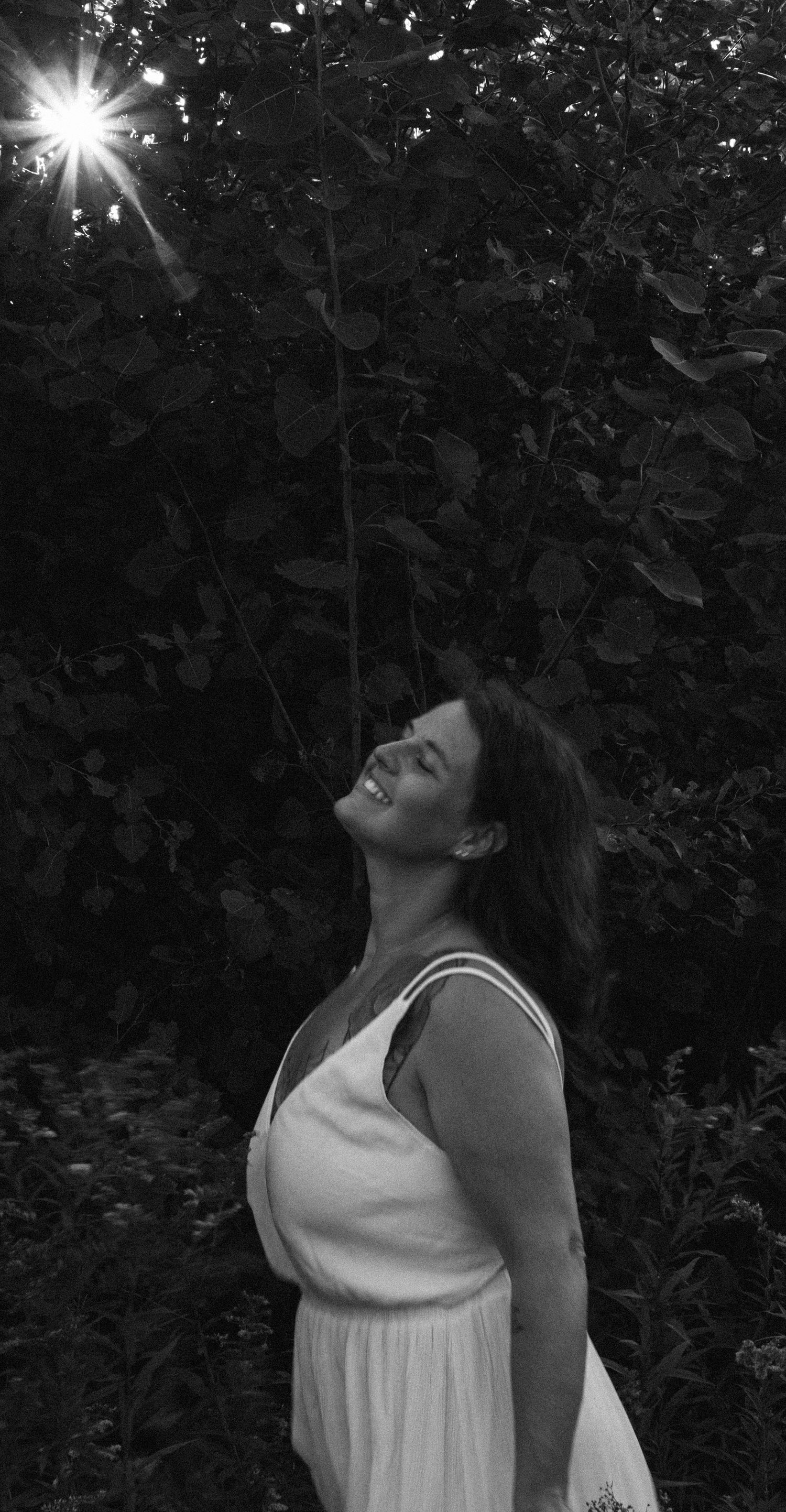 A woman in a white dress tilts her head back against a dark garden backdrop with sunbeams filtering through.