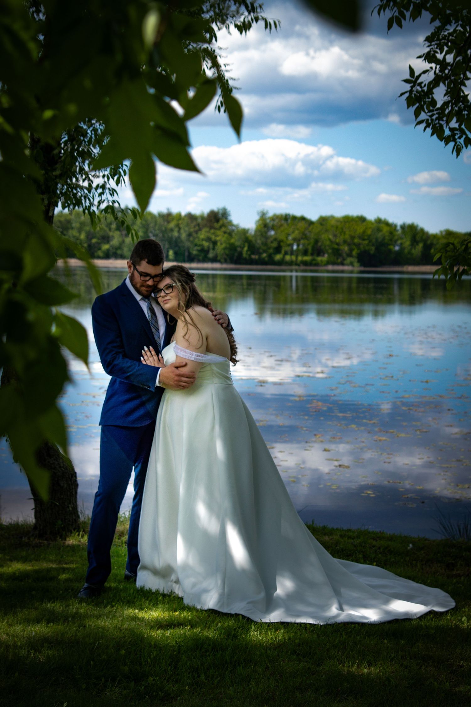 A romantic lakeside wedding photo featuring a couple embracing on a grassy shore with trees framing the peaceful water view.