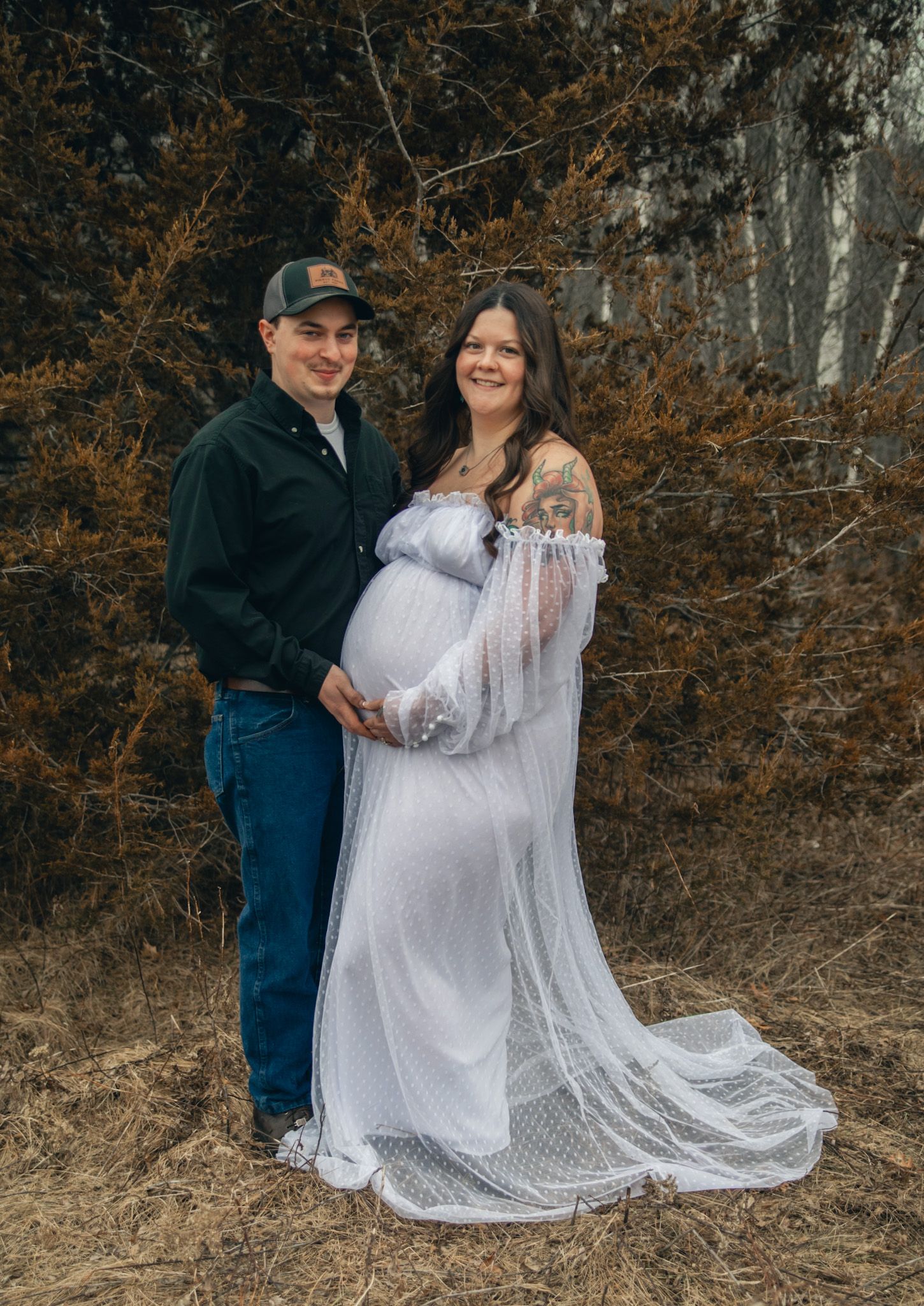 Couple poses outdoors for maternity photo, woman in white lace gown showing baby bump, man in cap and dark shirt.