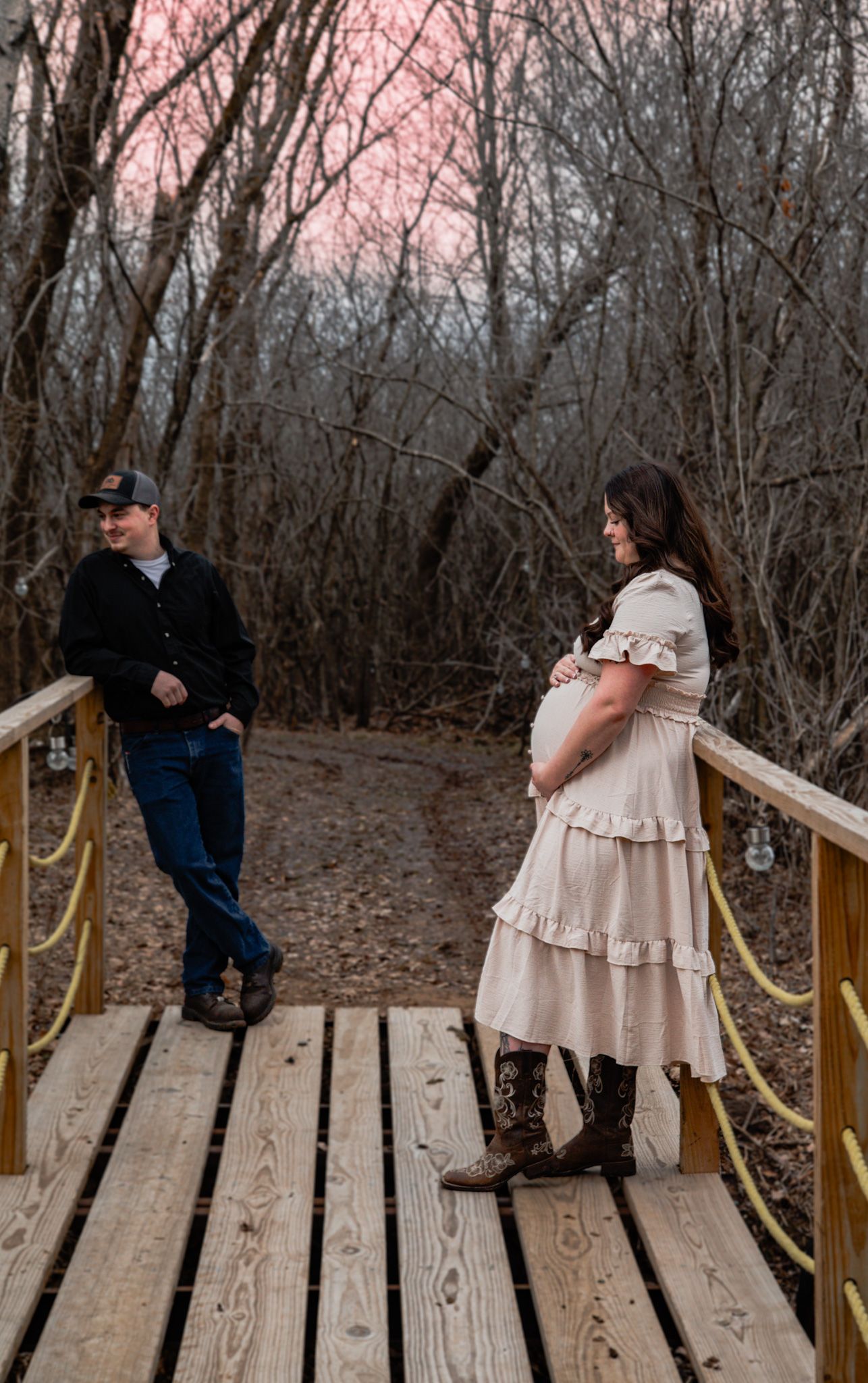 Expecting couple on wooden bridge at sunset, woman in beige dress and cowboy boots holding baby bump.