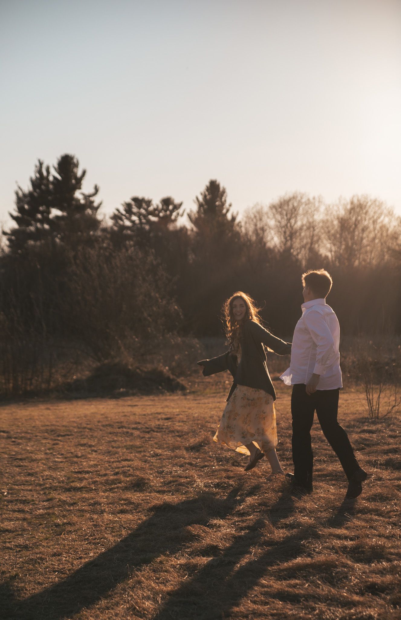 Couple walking together in sunlit field at dusk, surrounded by bare trees in warm golden light.