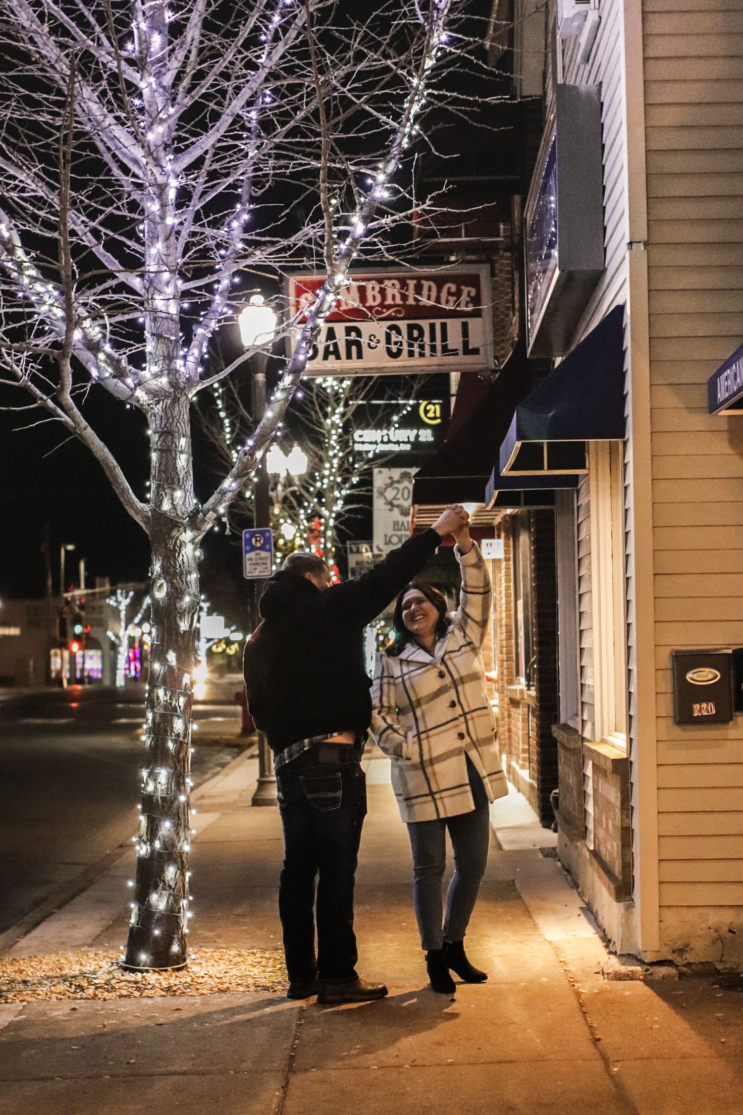 People stand and chat on a winter evening sidewalk illuminated by decorative white lights wrapped around bare trees.