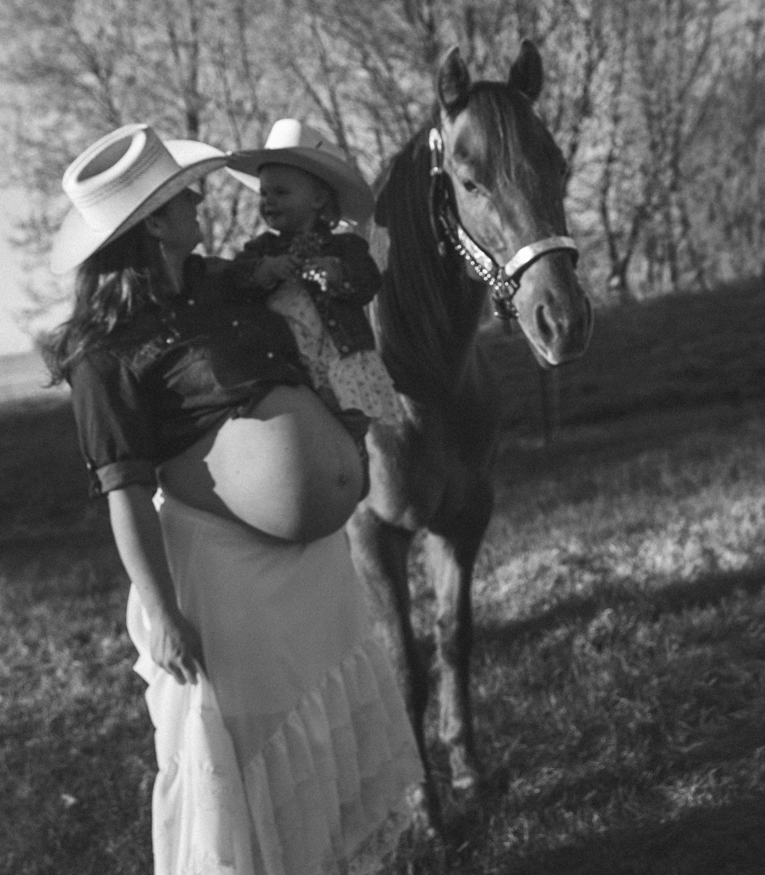 Black and white maternity photo of pregnant woman in cowboy hat and white skirt posing with a horse outdoors.