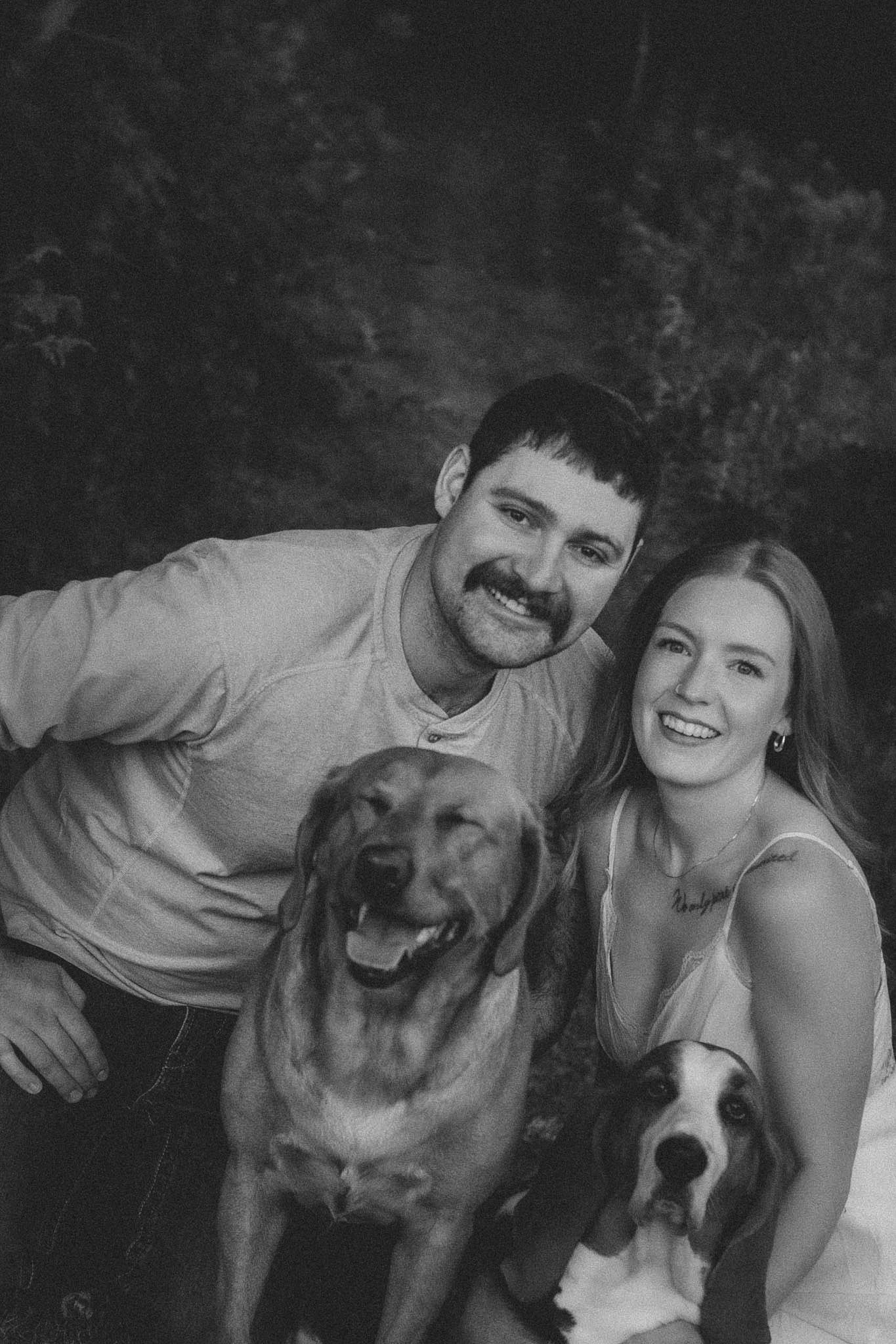 Black and white photo of a smiling couple posing outdoors with their two happy dogs, a Golden Retriever and a Beagle.
