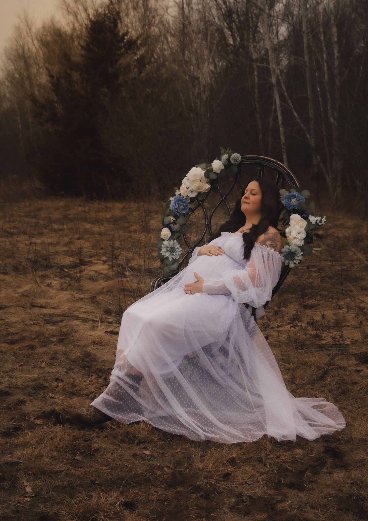 Maternity portrait of woman in sheer white dress seated in a flower-adorned arch chair outdoors.