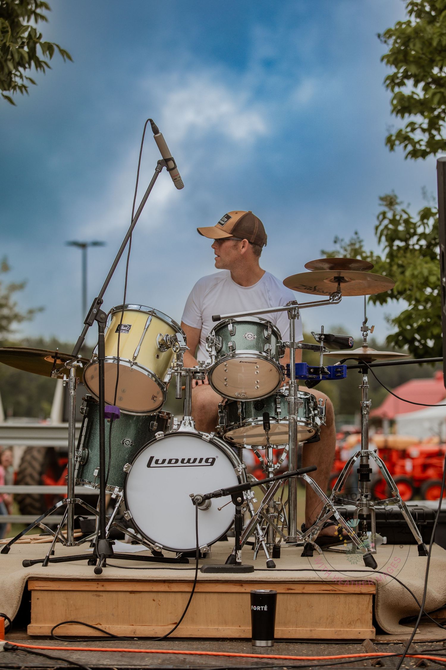 Drum kit set up on wooden platform outdoors under blue sky with trees in background.