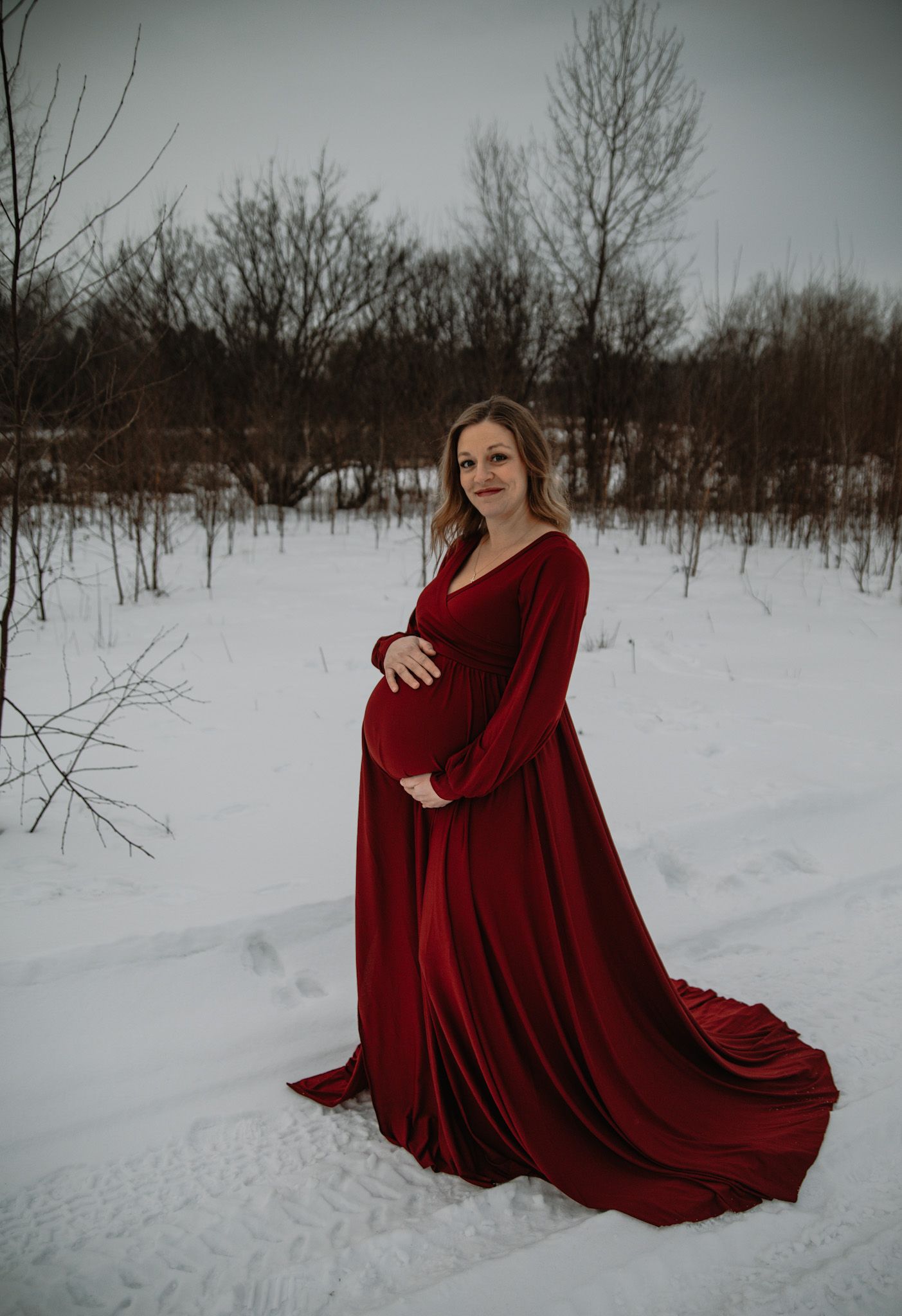 Expectant mother in flowing red velvet gown poses gracefully in snowy winter landscape with bare trees.