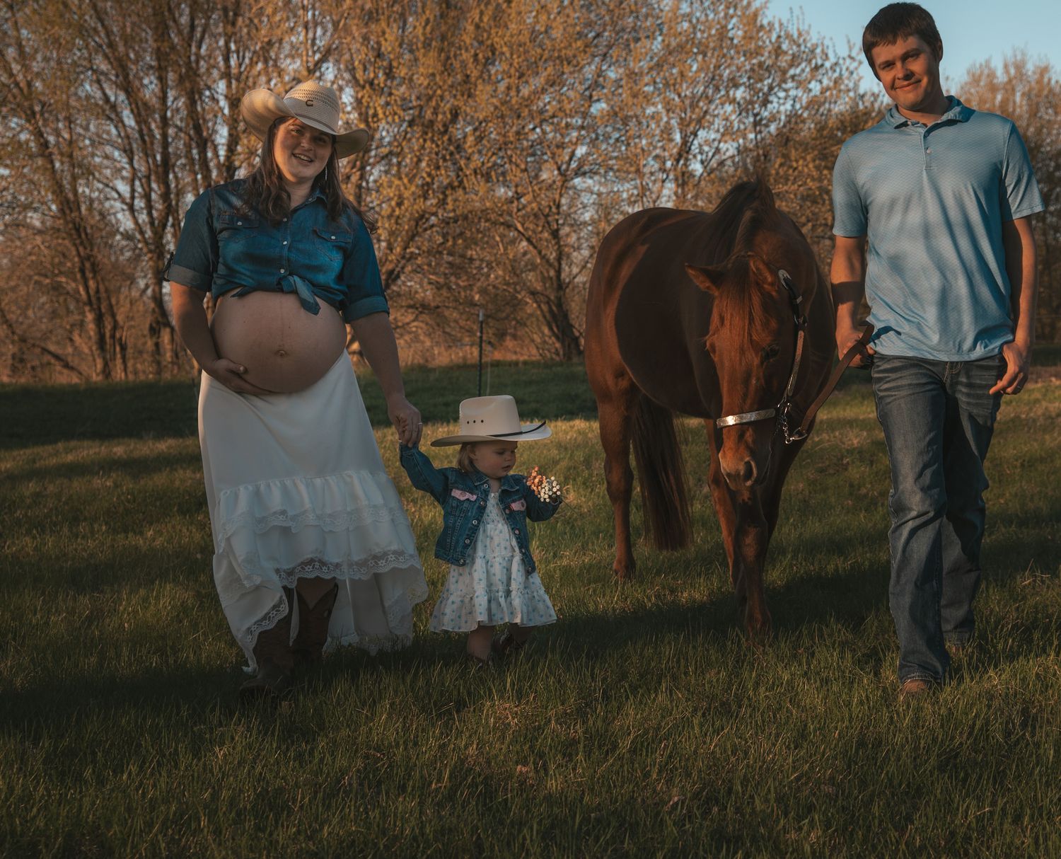 Pregnant woman and man with toddler in cowboy hats walking with a brown horse in a golden sunset field.